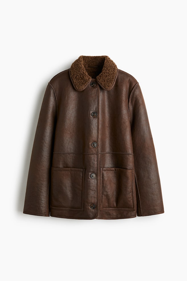 Tummanruskea keinonahkatakki, joka on kuvattu edestä, sisältää teksturoidun ruskean shearling-tyylisen kauluksen, edessä olevan nappikiinnityksen neljällä tummalla napilla ja kaksi suurta paikkataskua alhaalla edessä. - 1