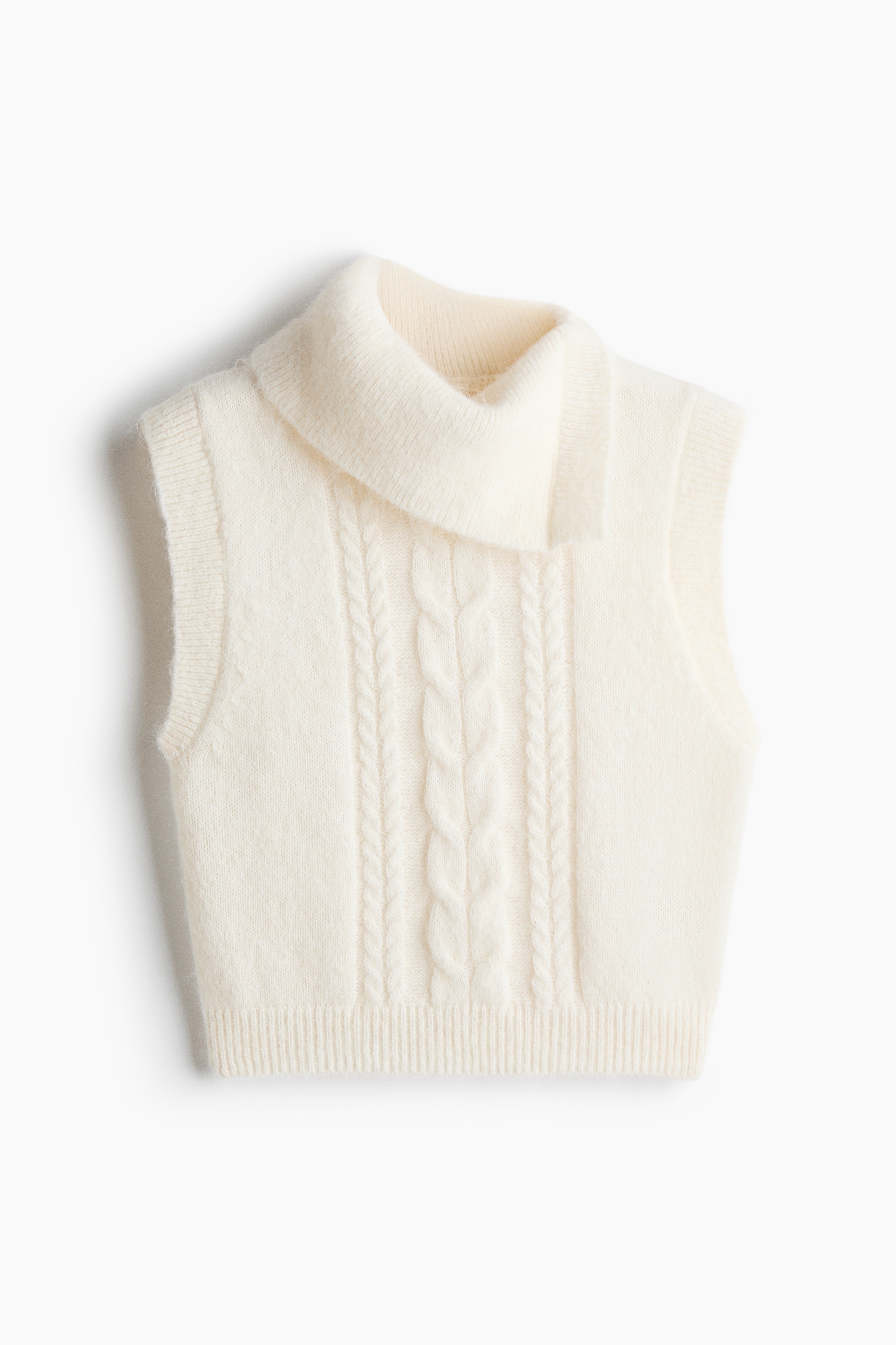 Người mẫu đang mặc một chiếc áo vest len màu kem với phần thân trước đan họa tiết cable-knit, cổ gập lệch bất đối xứng và gấu áo bo gân. Chiếc áo vest dáng ngắn được phối cùng quần jeans xanh ống rộng và thắt lưng đen.