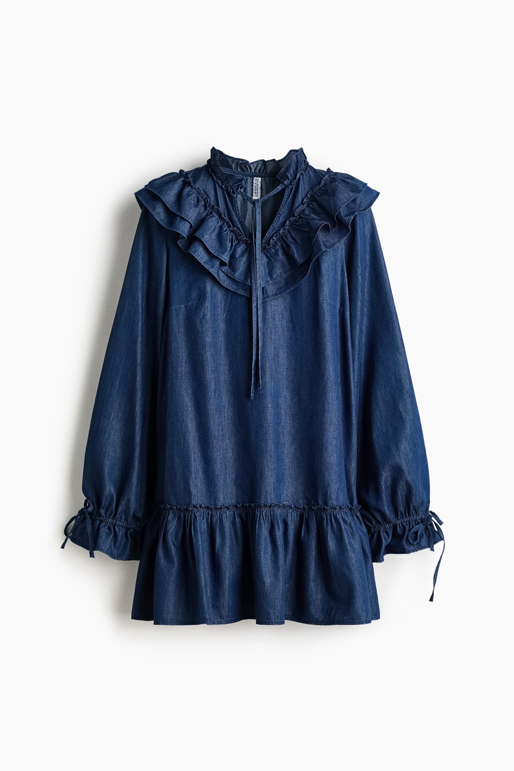 Ampliar la imagen: Un vestido de mezclilla azul oscuro está colgado, con un cuello alto con volantes, una abertura en forma de V con lazos, y grandes volantes que se extienden por el pecho y los hombros. El vestido presenta mangas largas con puños fruncidos con detalle de lazos y un dobladillo con volantes.