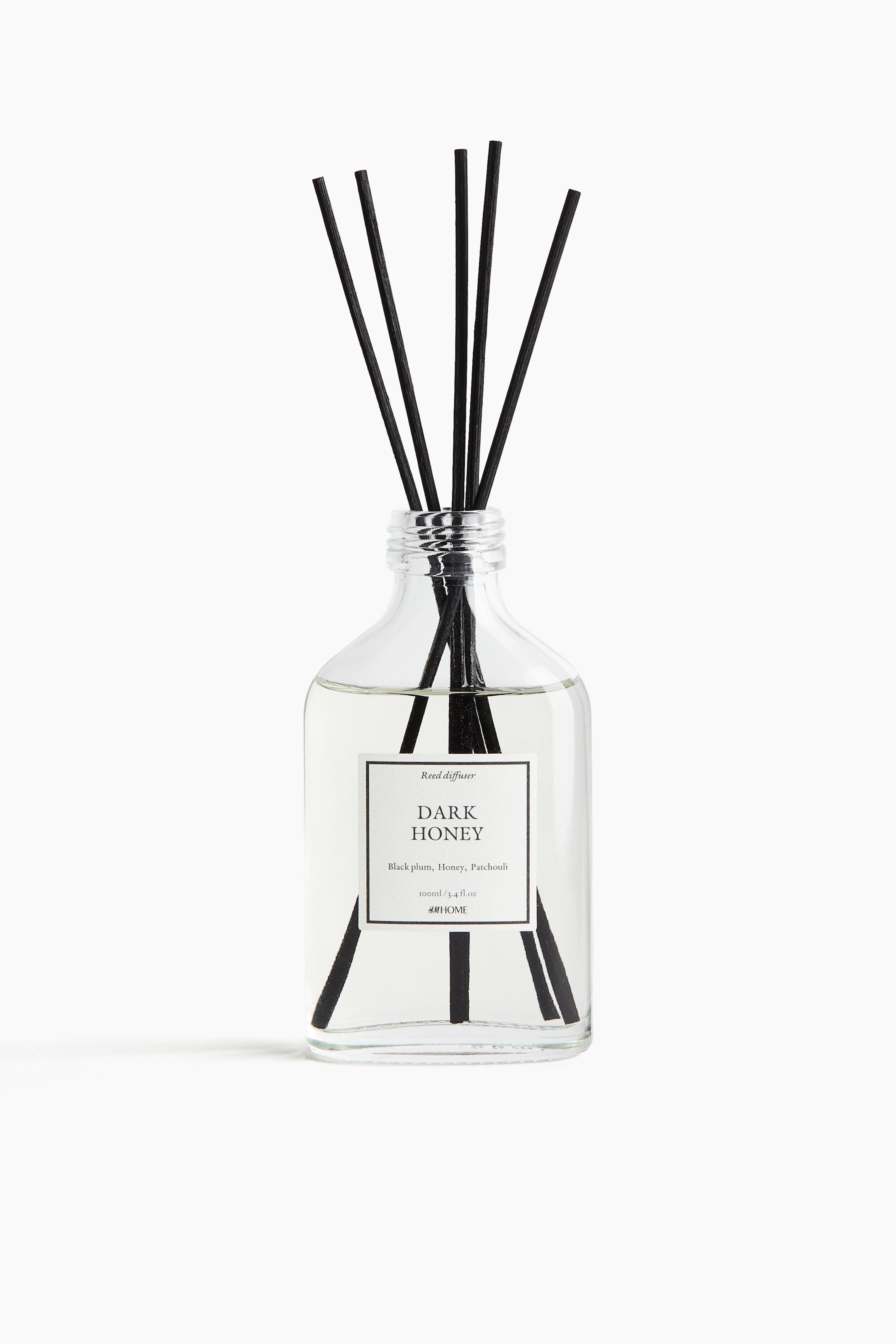 View larger image: A reed diffuser is presented from the front, featuring a clear, square-shaped glass bottle with rounded shoulders, a ribbed neck, and a white label reading "DARK HONEY" on its front. Light-colored liquid is filling the bottle, and several black reeds are extending from the neck and submerged in the liquid.