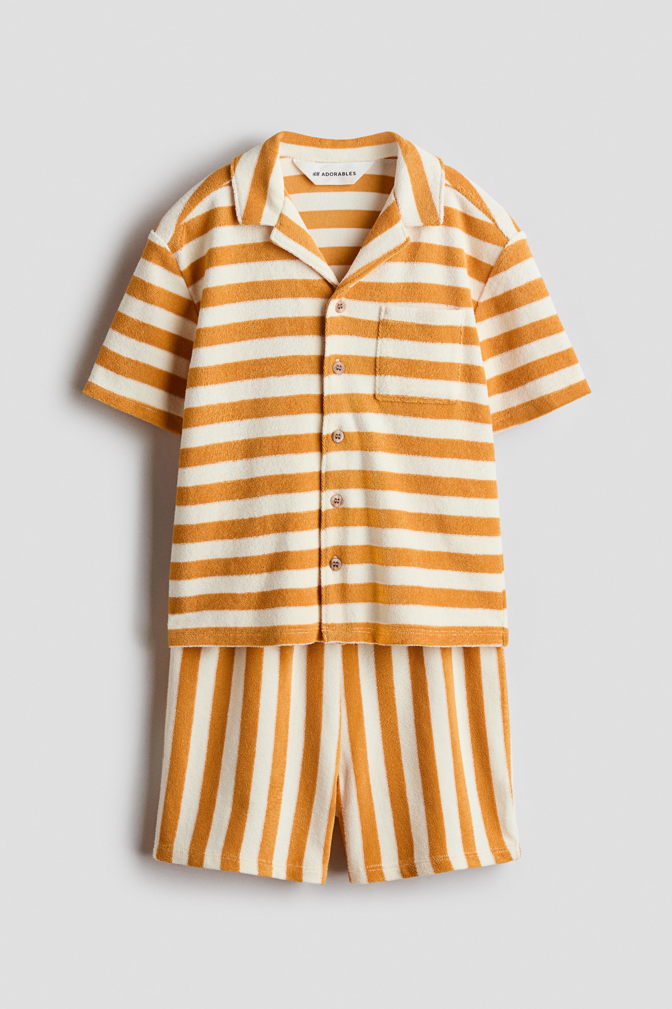 Grotere afbeelding bekijken: Een badstof overhemd met korte mouwen en bijpassende shorts set wordt van voren weergegeven, beide met oranje en crèmekleurige strepen. Het overhemd is ontworpen met een resortkraag, bruine knopen aan de voorkant, een borstzak met klep en horizontale strepen, terwijl de shorts bijpassende verticale strepen hebben.