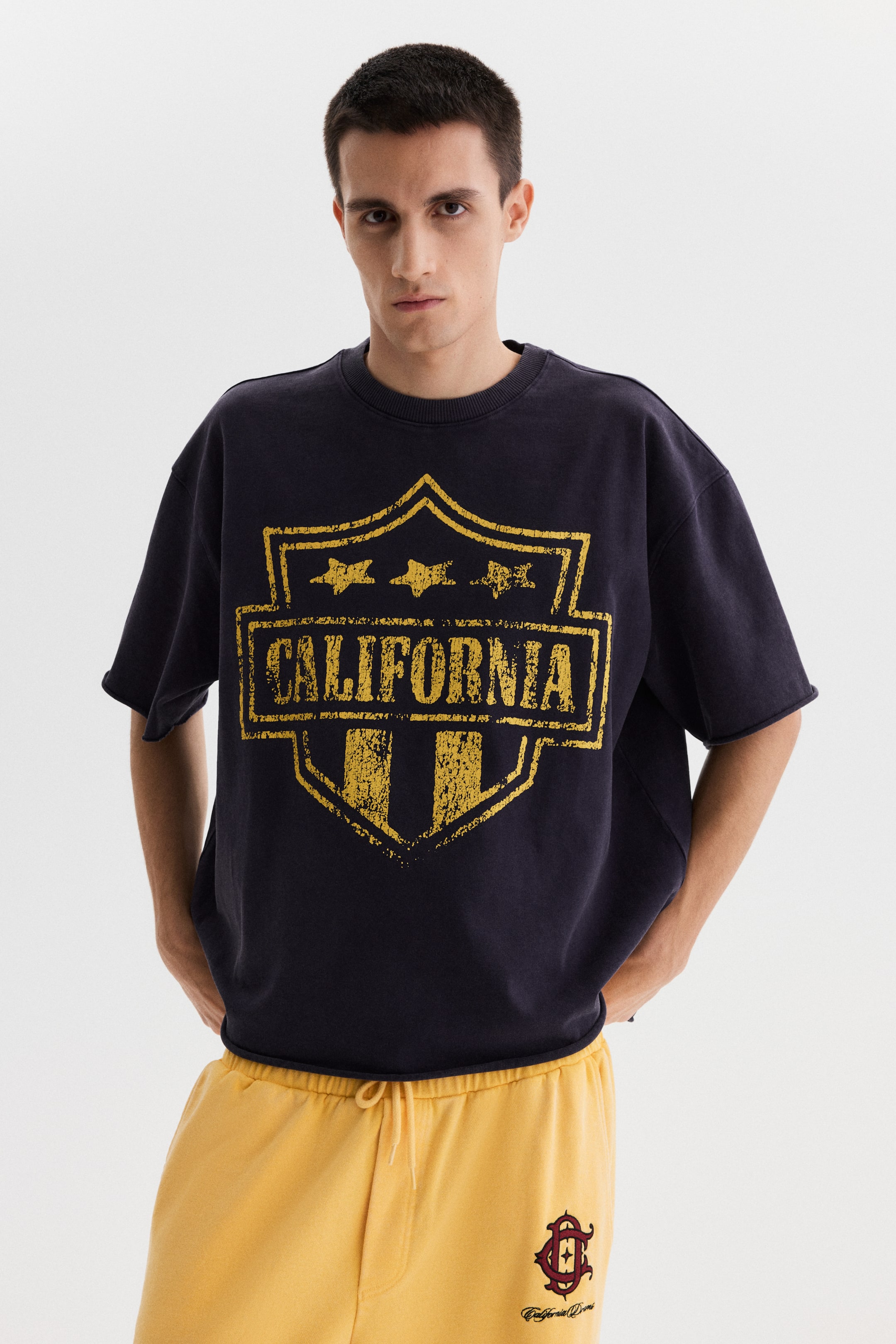View larger image: A dark blue short-sleeve sweatshirt features a large, distressed yellow shield graphic on the front, displaying "CALIFORNIA" on a banner with three stars above. The sweatshirt has an oversized fit with a ribbed crewneck and raw-edged short sleeves and hem.