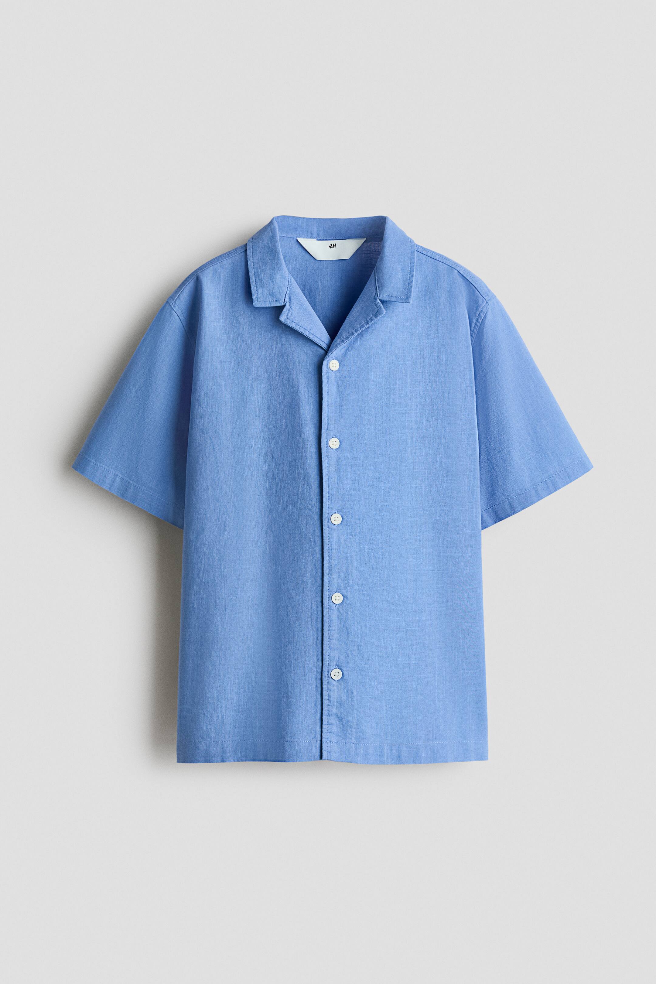View larger image: A blue short-sleeved shirt is on display, showcasing its front with a camp collar and a full button-front closure adorned with white buttons. The fabric exhibits a subtle texture.