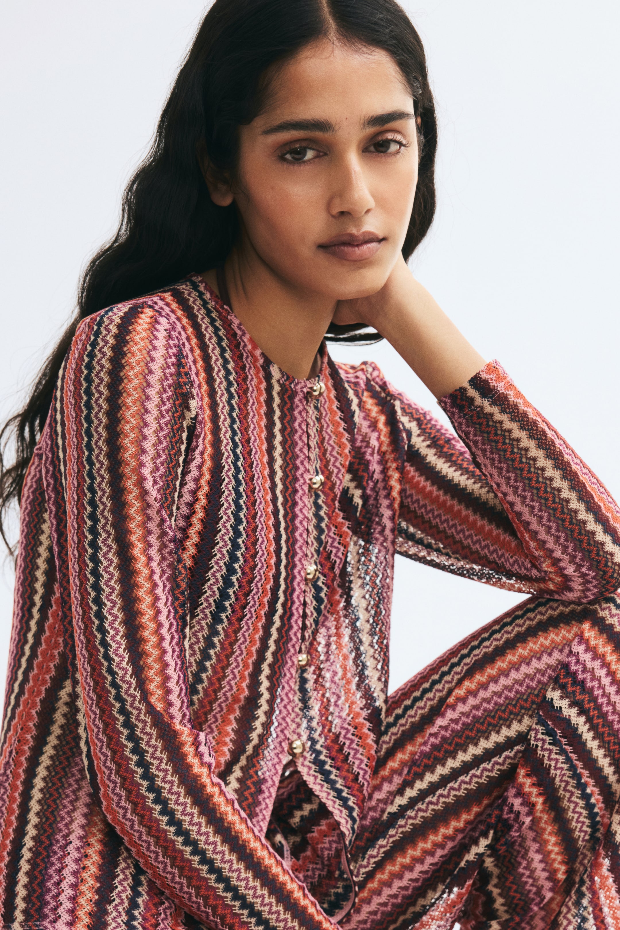 View larger image: A fitted cardigan in textured jersey features a multicoloured zigzag stripe pattern in pink, orange, and brown. The cardigan has a round neckline, long sleeves, and gold-tone dome buttons down the front, and is paired with matching trousers.