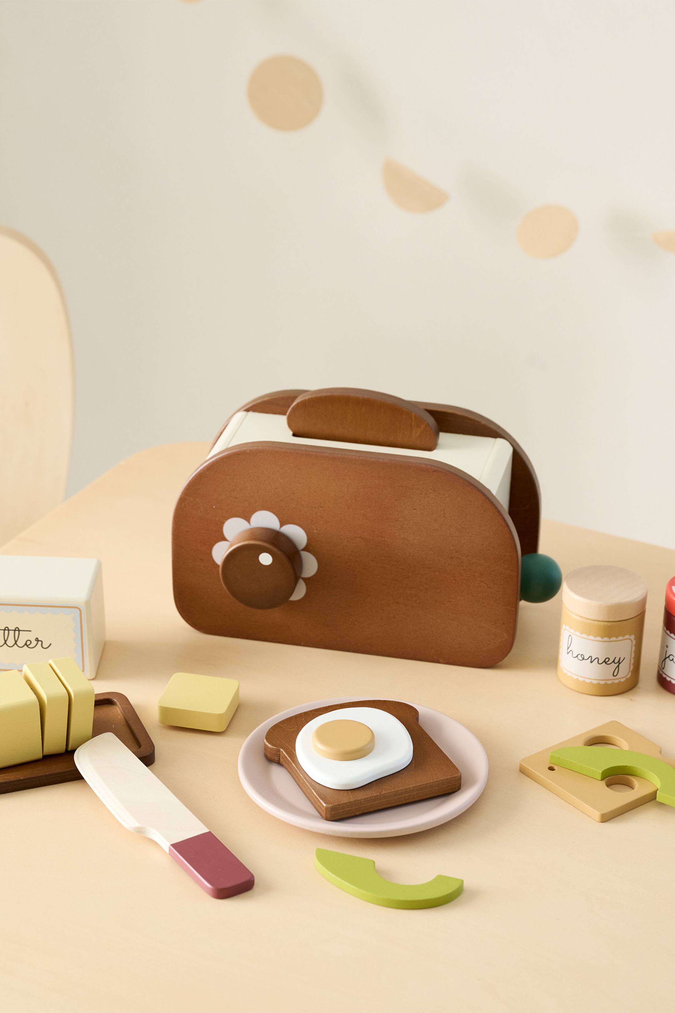 Grotere afbeelding bekijken: Een houten speelgoedbroodrooster staat onder een lichte hoek, waarbij de bruine buitenkant en twee crèmekleurige sleuven voor toast worden getoond. Twee stukken houten toast komen uit de sleuven tevoorschijn, terwijl een bruine bedieningsknop met een wit madeliefjesontwerp zichtbaar is aan de zijkant, samen met een groene hendel.