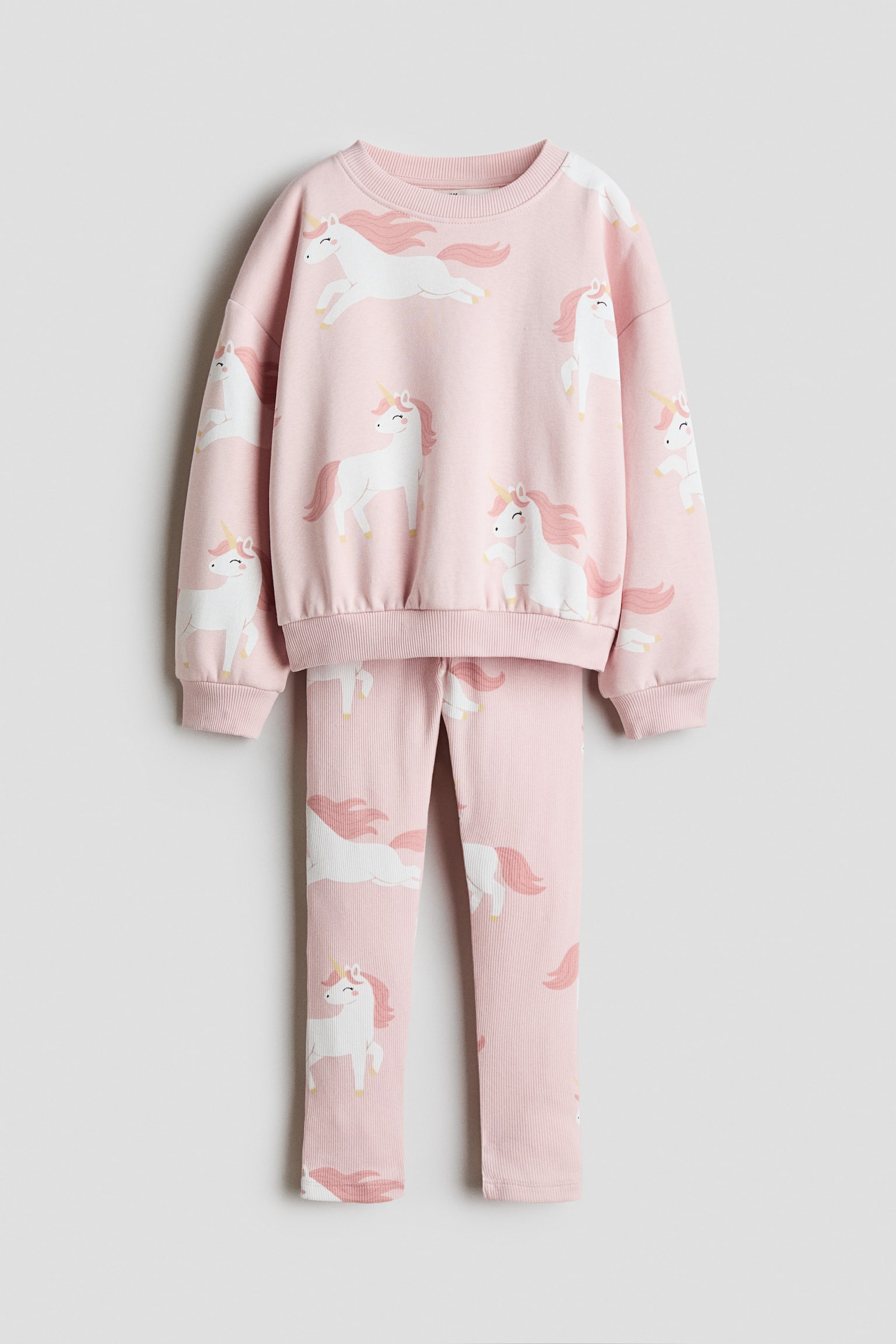Ein zweiteiliges Set in Rosa wird von vorne gezeigt und besteht aus einem langÃ¤rmeligen Pullover und einer passenden Leggings mit einem Allover-Einhorn-Print in WeiÃ und Rosa. Der Pullover ist mit einem gerippten Rundhalsausschnitt, Ã¼berschnittenen Schultern sowie gerippten BÃ¼ndchen und Saum zu sehen, wÃ¤hrend die abgestimmte Leggings gerade darunter hÃ¤ngt.