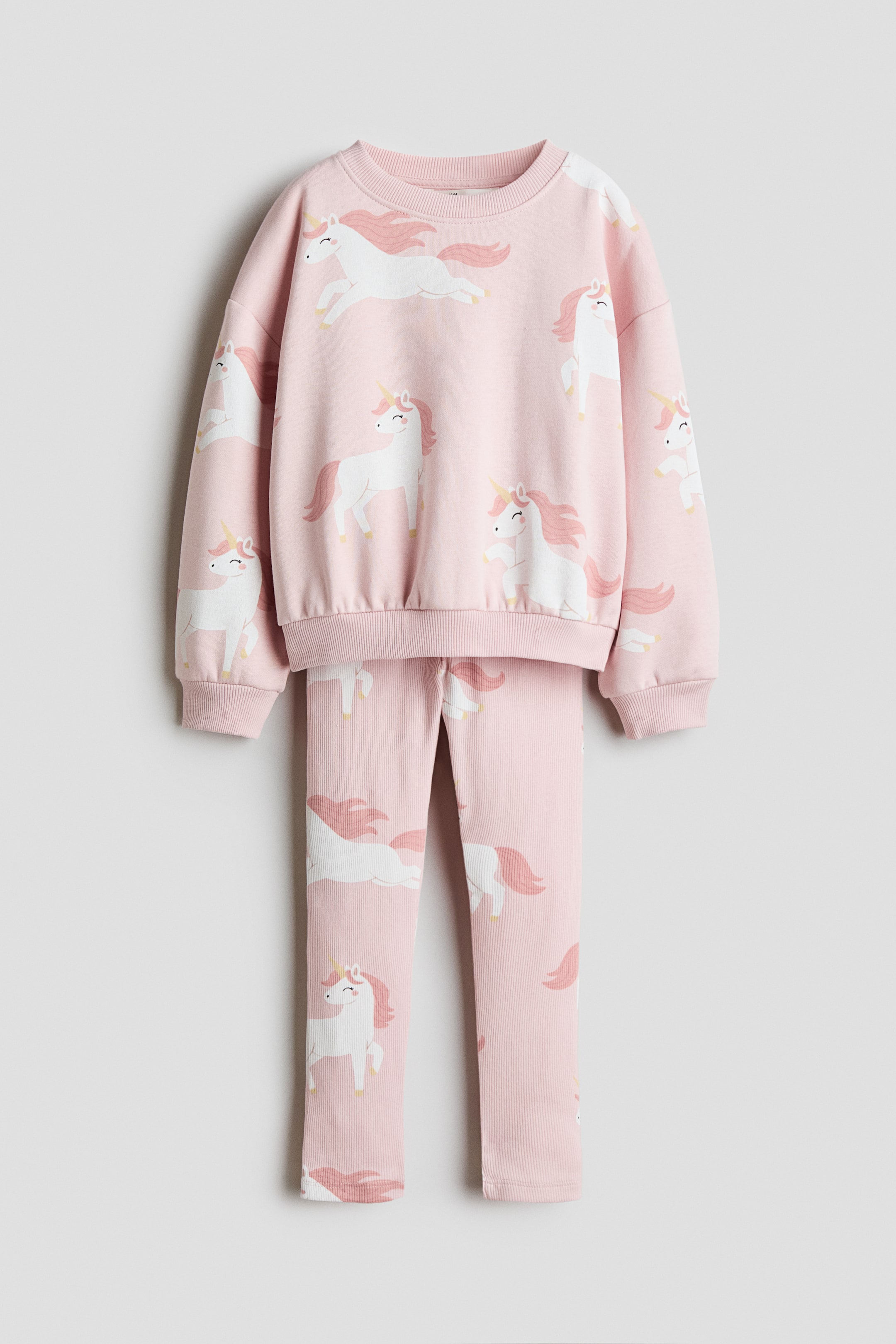 View larger image: A pink two-piece set is displayed from the front, featuring a long-sleeved sweater and matching leggings with an all-over white and pink unicorn print. The sweater shows a rib-trimmed round neckline, dropped shoulders, and ribbed cuffs and hem, while the coordinating leggings hang straight underneath.