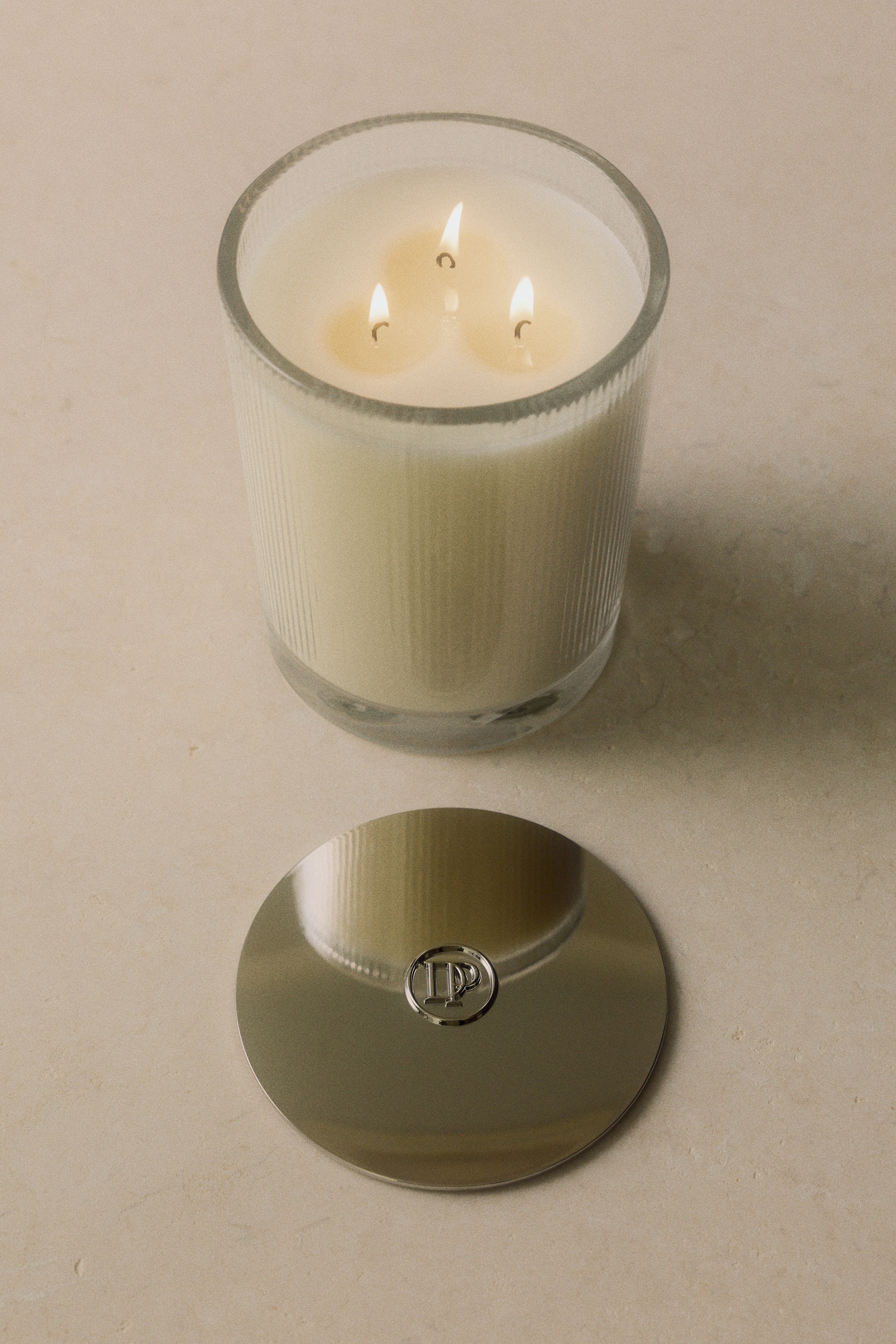 Una candela profumata in un barattolo di vetro trasparente rigato, riempito di cera color crema e sormontato da un coperchio argentato lucido, Ã¨ posizionata accanto alla sua confezione bianca minimalista. Sul fronte della scatola, nella parte superiore, Ã¨ presente la scritta in rilievo "RICH AMBER".