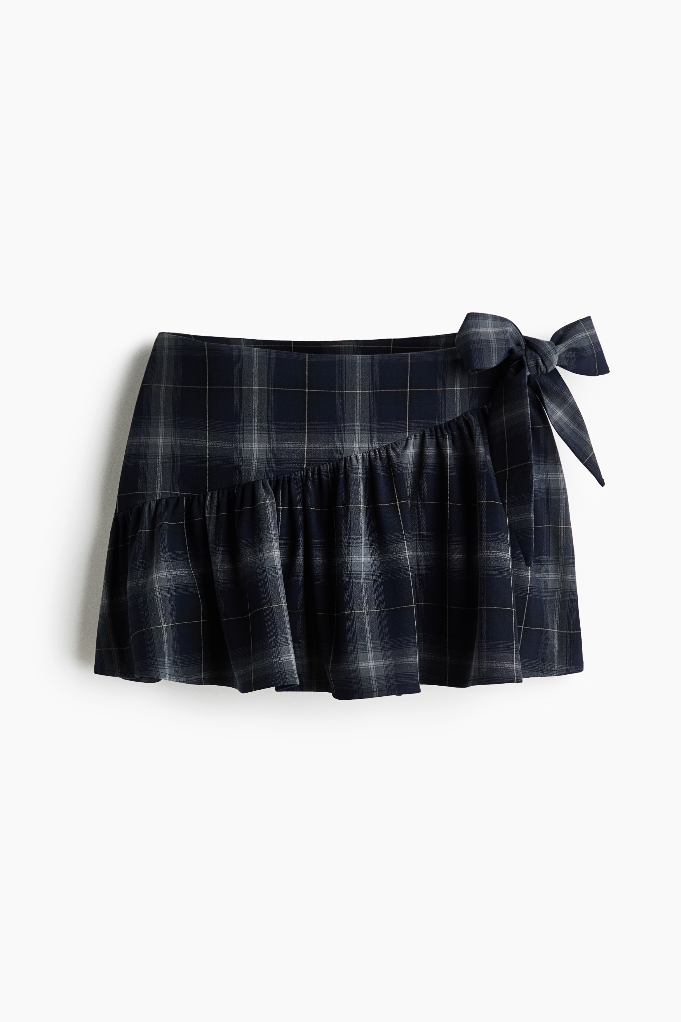 A dark plaid mini skort featuring an asymmetrical gathered seam and a long, wide fabric tie at the side is worn by the model. The skort is styled with a dark blue long-sleeve sweater, white socks, and blue sneakers.