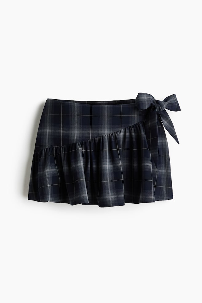 A plaid mini skirt is featuring a navy blue, gray, and white check pattern, a gathered ruffle detail diagonally across the front, and a decorative bow tied on the upper right side. - 1