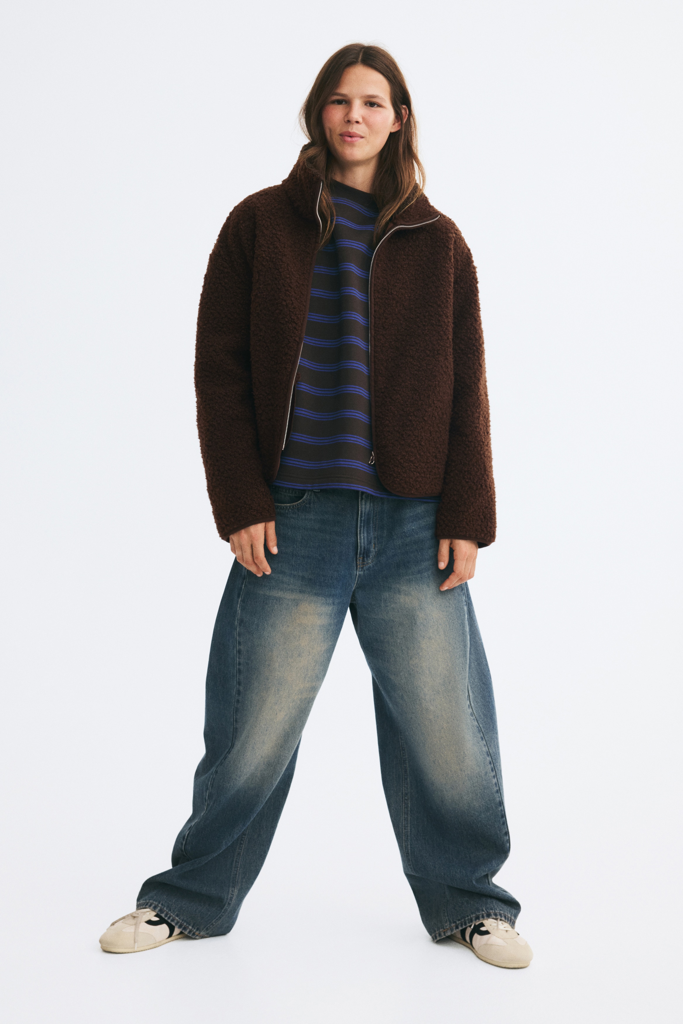 A dark brown fleece jacket is shown from the front, featuring a full-length zipper with a silver pull, a high stand-up collar, and two subtle side pockets. A brown trim is visible along the jacketâs curved bottom hem.