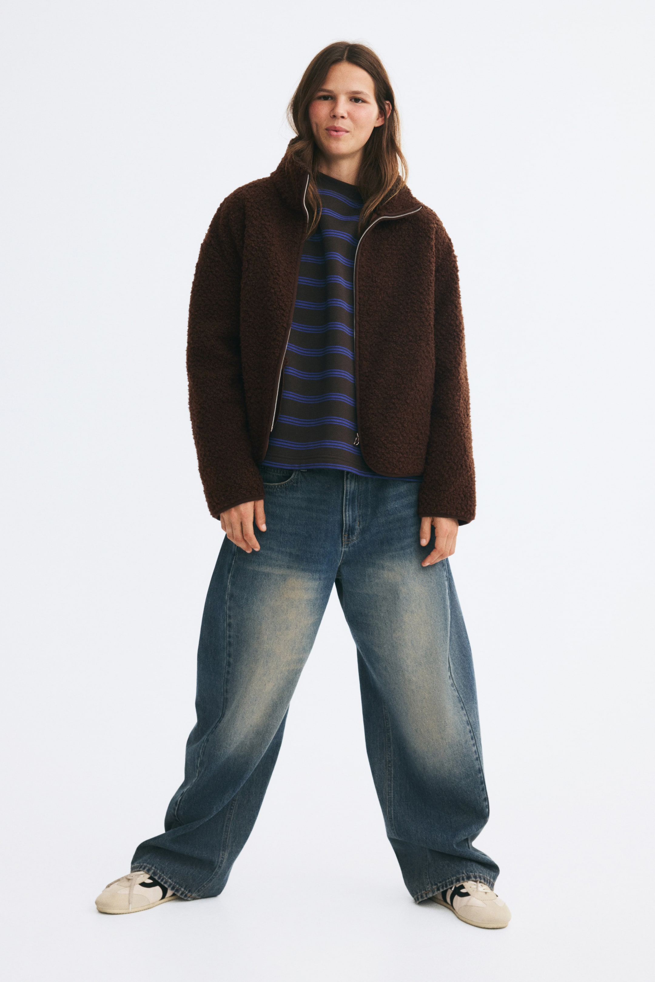 View larger image: A dark brown teddy fleece jacket with a stand-up collar and front zipper is worn open by the model. The jacket is layered over a striped long-sleeve shirt and paired with wide-leg blue jeans.