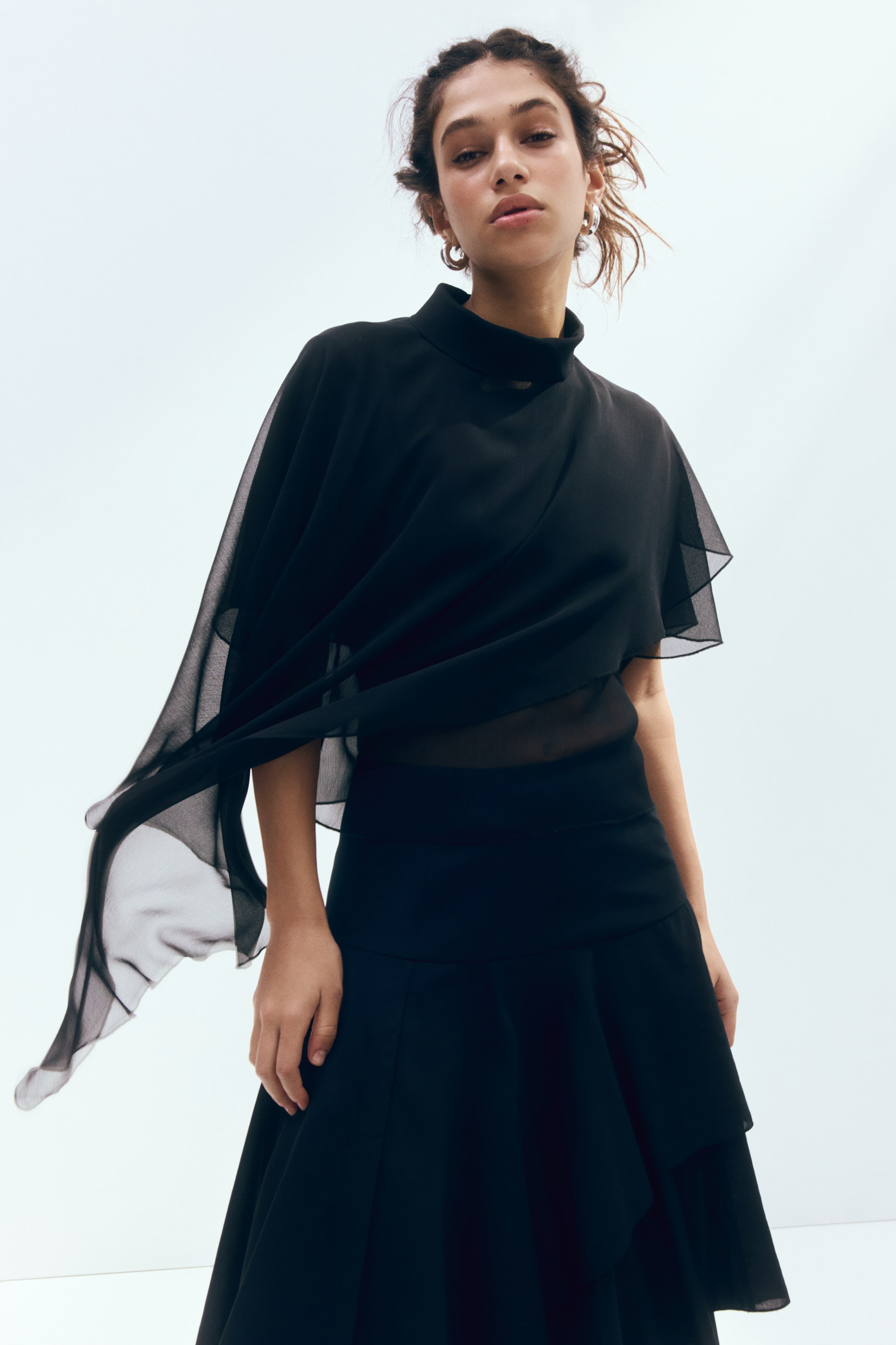 View larger image: A black top and skirt are displayed, angled slightly to the right. The top boasts a high neckline, a long, flowing sheer panel extending from the wearer's right shoulder, layered sheer fabric on the wearer's left shoulder, and a sheer mid-section. The skirt is designed with tiered ruffles.