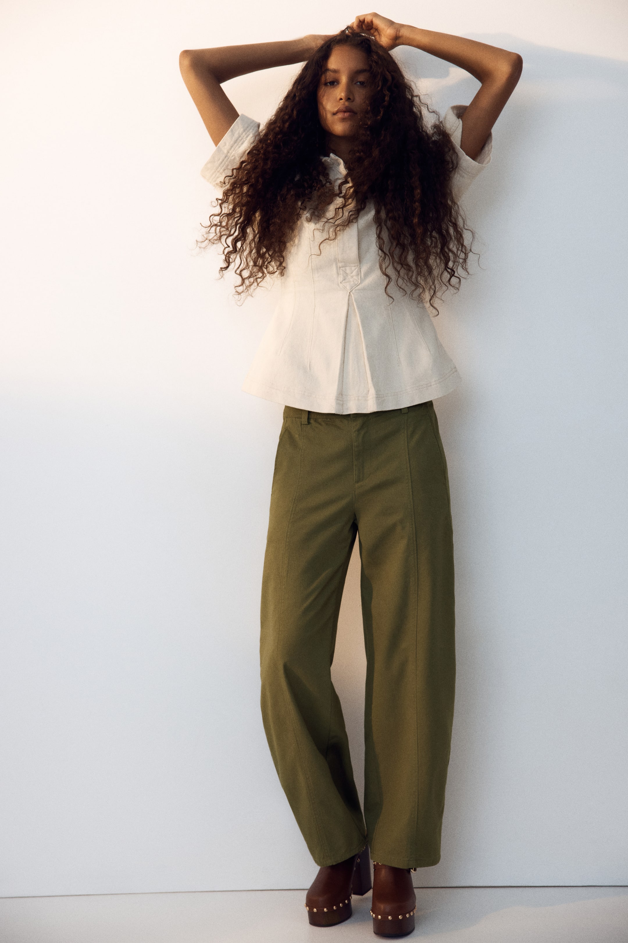 View larger image: An off-white textured short-sleeved top features cuffed sleeves, a stand collar, a buttoned placket, and pleated details that create a flared hem. Olive green wide-leg pants showcase a prominent front seam on each leg, and brown platform clogs with stud details complete the outfit.