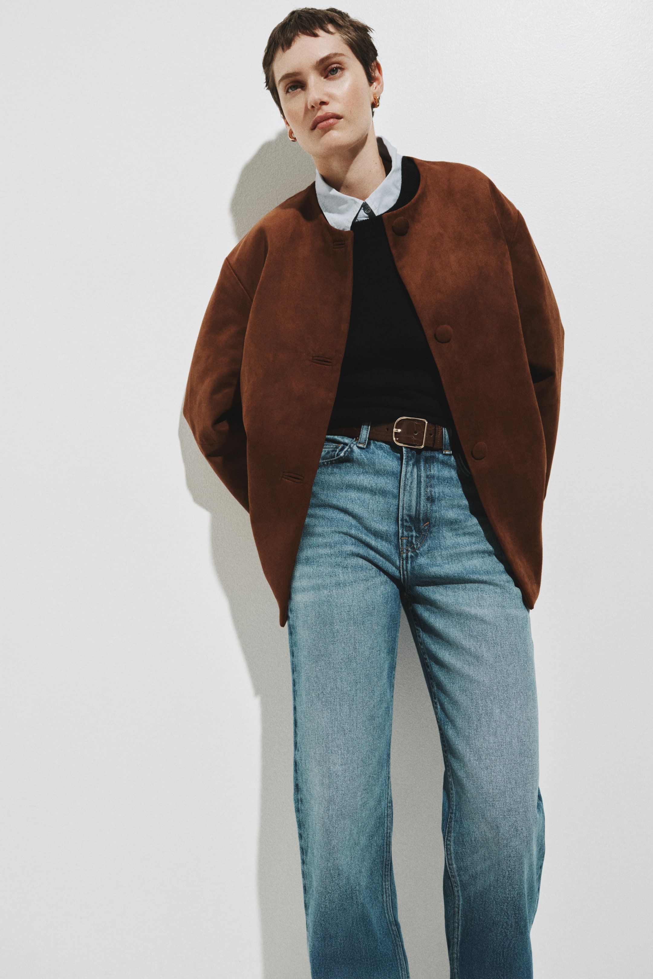 Visualizza immagine più grande: Giacca marrone dall'effetto suede, dal taglio rilassato e leggermente oversize, con design senza colletto e scollatura rotonda, con tre grandi bottoni circolari sul davanti e due tasche con pattina. La giacca è presentata aperta, lasciando intravedere un outfit composto da capi sovrapposti.