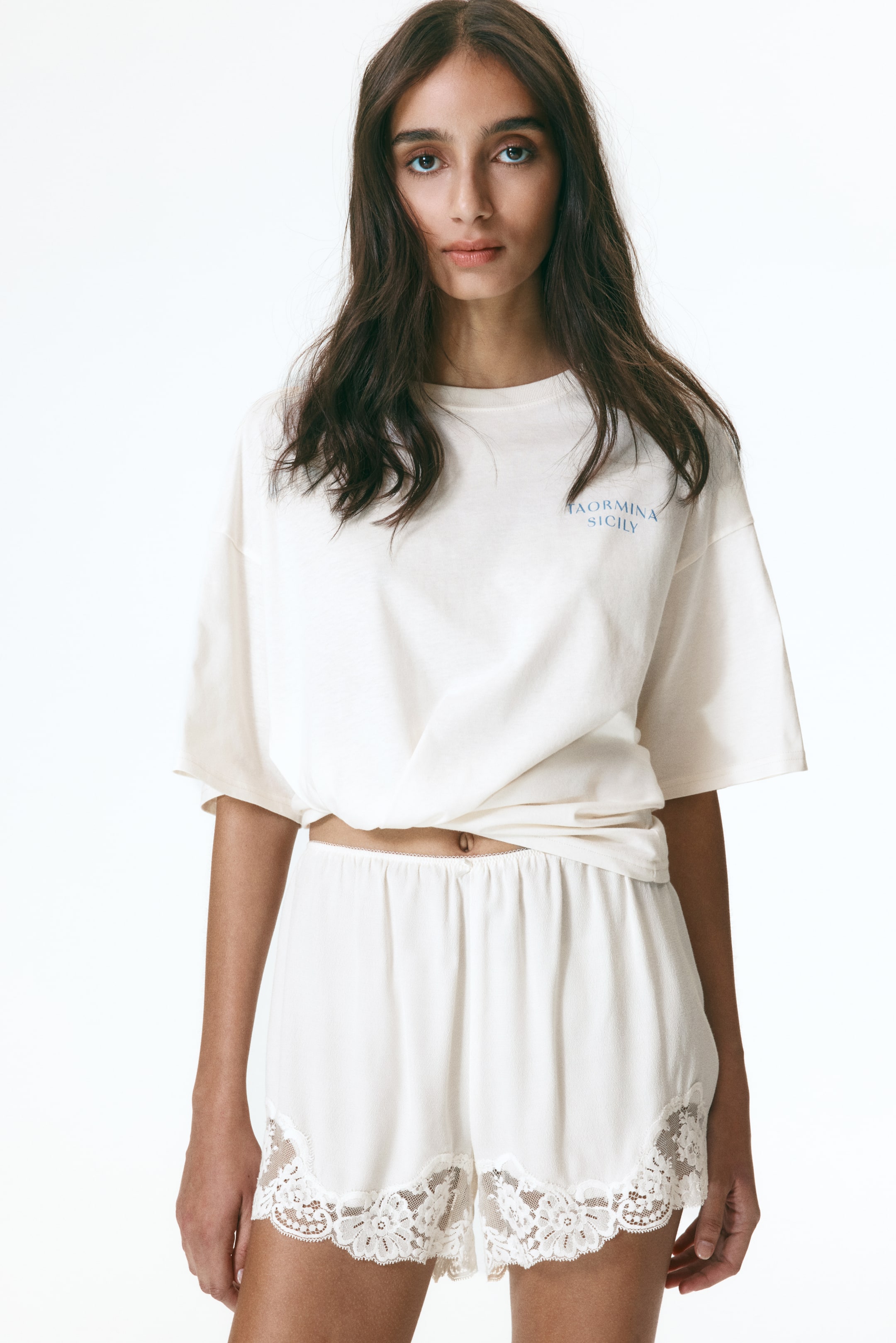 拡大画像を表示: オフホワイトのツーピースルームウェアセットが正面から紹介されています。リラックスフィットのTシャツは、クルーネック、ショートスリーブ、「TAORMINA SICILY」の文字が胸にあり、裾は前で結ばれています。マッチングショーツは、ウエストにゴムが入っており、脚の開口部には広いスカラップレーストリムが施されています。