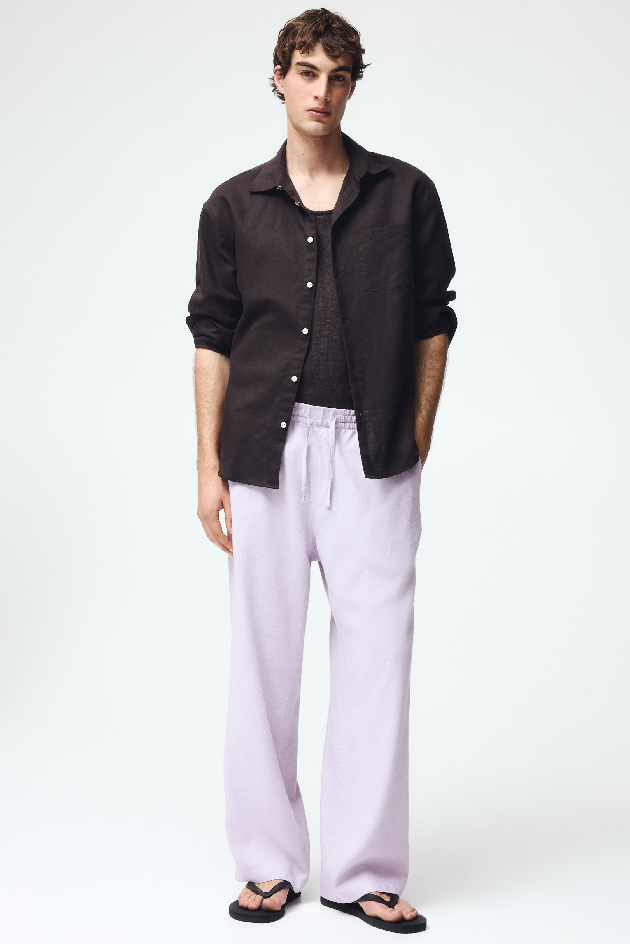 View larger image: Presenting a dark brown linen shirt, showcasing a spread collar, white buttons, and a single left chest pocket, worn open over a dark undershirt with its sleeves rolled up. This is paired with light purple wide-leg pants featuring an elasticated drawstring waist, complemented by black flip-flops.