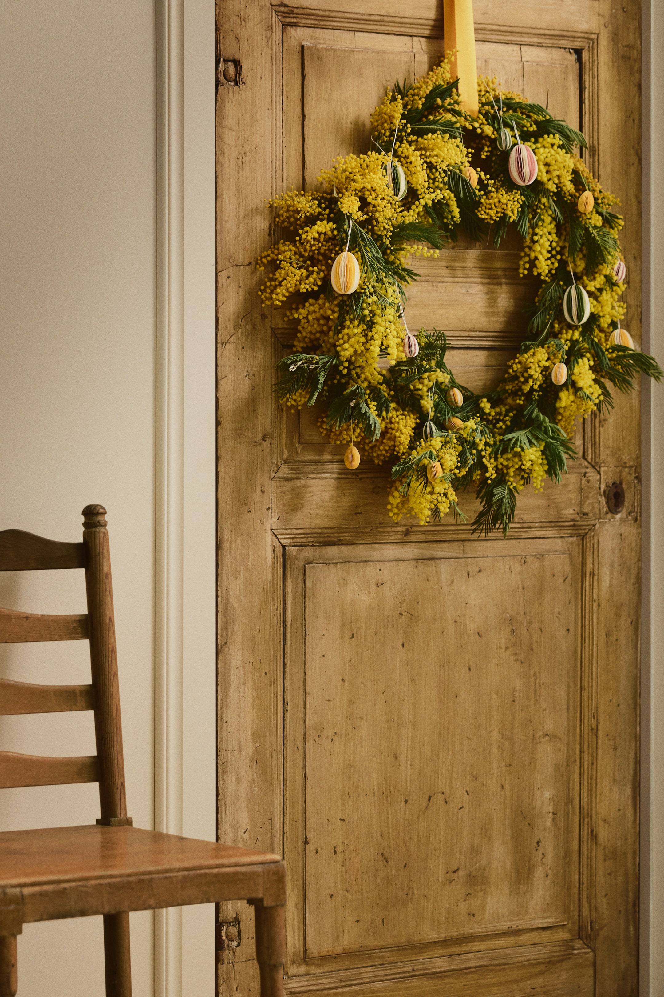 Grotere afbeelding bekijken: Een krans met dichte trossen van felgele mimosa bloemen en donkergroen loof. Verschillende gestreepte, ei-vormige ornamenten hangen aan dunne touwtjes van de krans, en een gele lint houdt het aan de bovenkant vast.