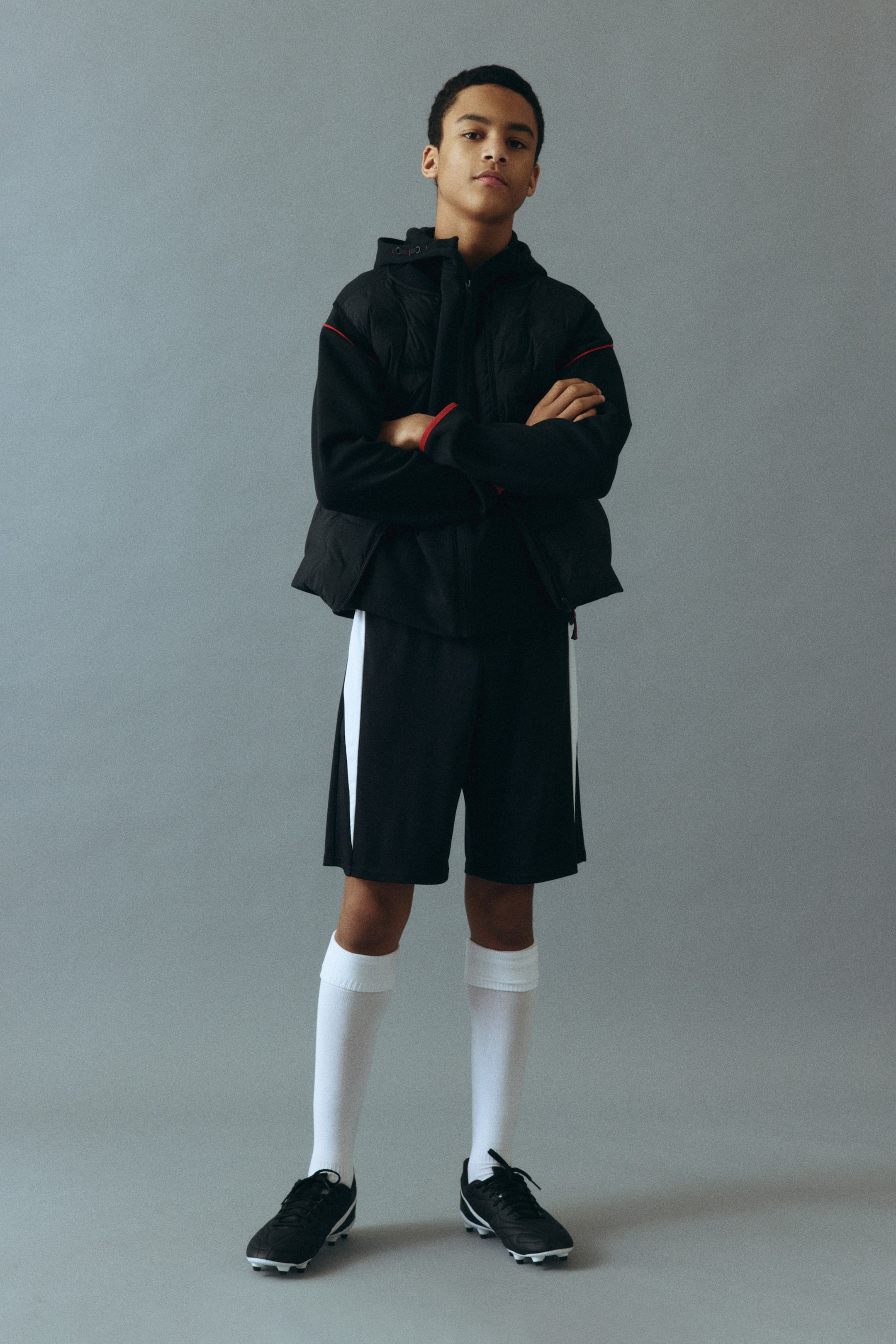 Näytä suurempi kuva: Musta hupullinen takki, jossa on tikattu vartalo ja kontrastiväriset pitkät hihat, joiden hihansuissa on punaista reunusta. Mustissa shortseissa on valkoinen pystysuora raita kummallakin sivulla. Valkoiset polvisukat peittävät sääret, ja mustat urheilukengät viimeistelevät ilmeen.