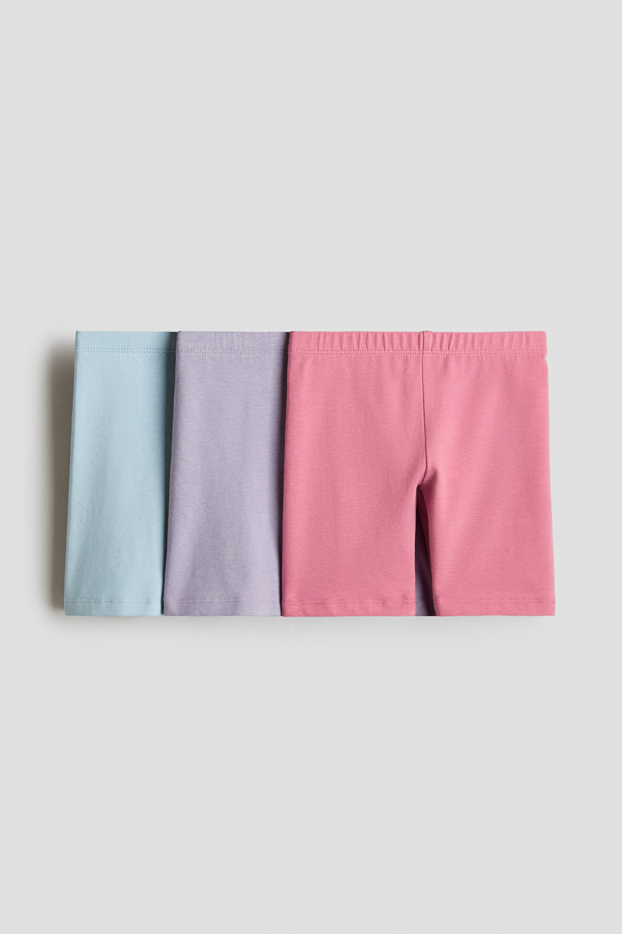 View larger image: Cycling shorts are presented as three pairs, each made of soft cotton jersey and featuring an elasticated waist. The shorts appear in light blue, light purple, and pink, overlapping slightly from left to right.