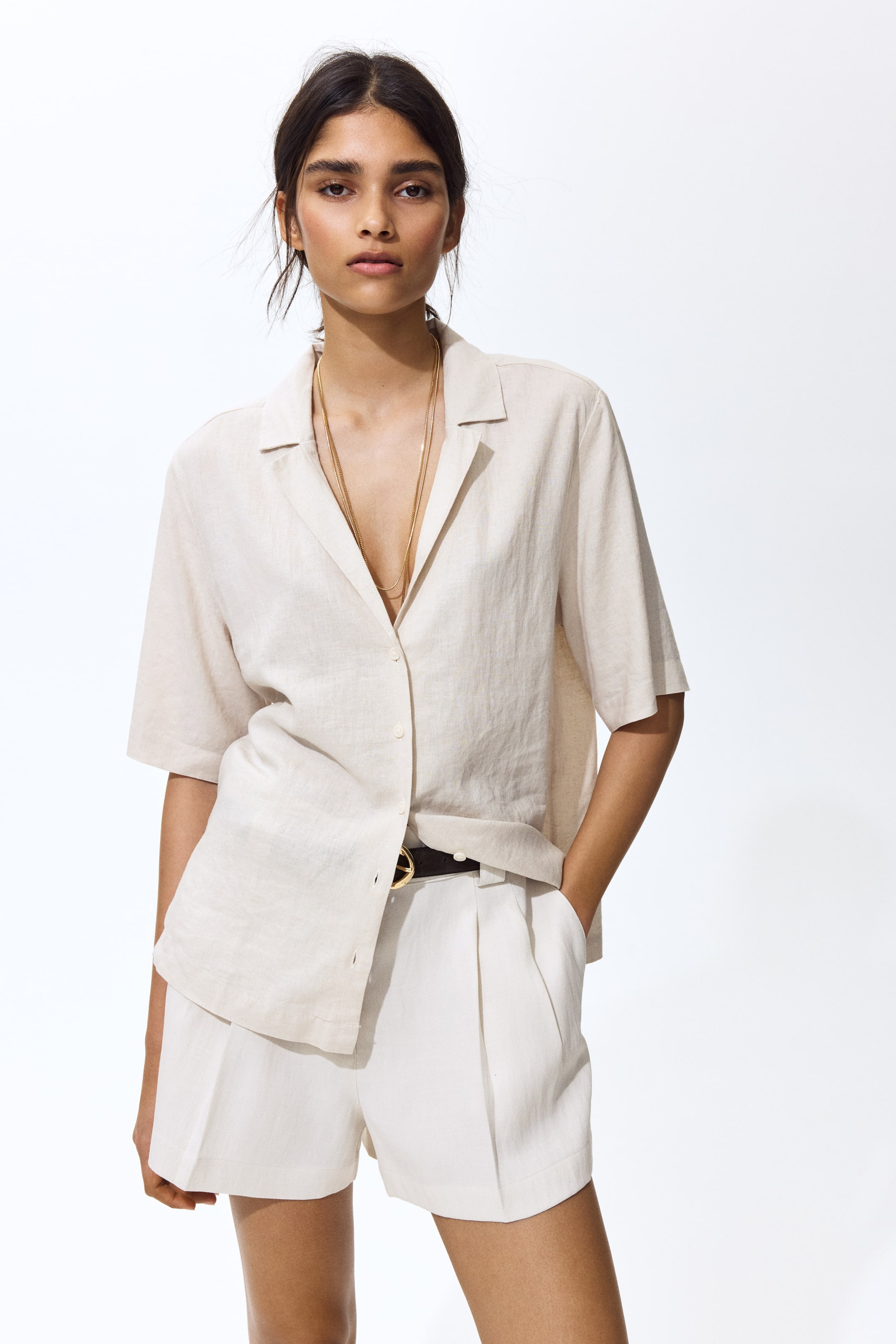 Vezi imaginea mai mare: O cămașă din in bej deschis, cu mâneci scurte și guler revere, cu nasturi în față, este purtată, parțial băgată în pantaloni scurți plisati, de culoare alb murdar, asortată. Pantalonii scurți sunt strânși cu o curea închisă la culoare, cu o cataramă aurie, iar la gât sunt vizibile coliere stratificate cu tonuri aurii.