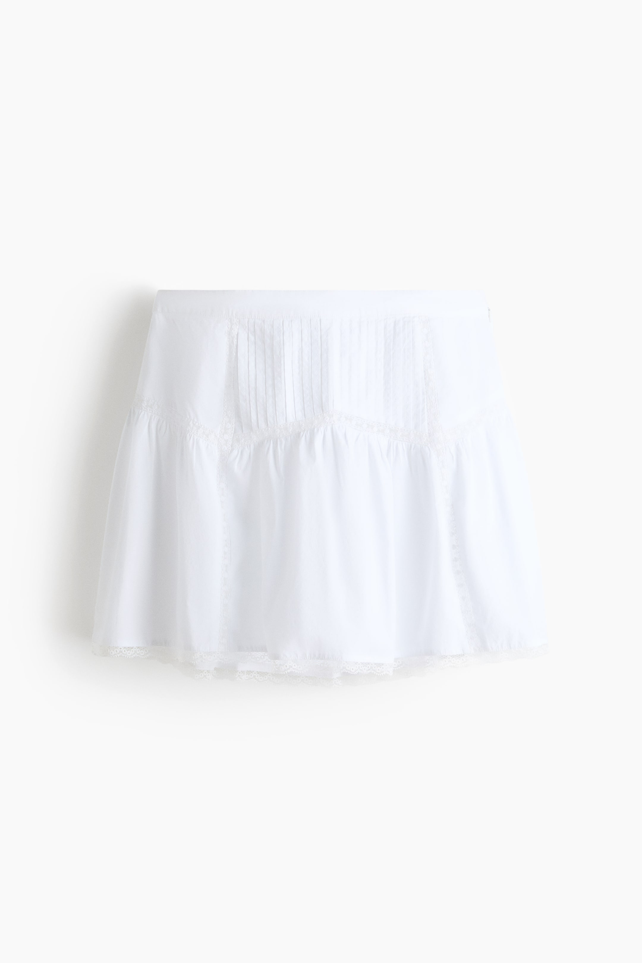 View larger image: The white mini skirt showcases its front, adorned with vertical pintucks and lace inserts on the upper portion. A horizontal lace trim elegantly separates this from a gathered lower tier, which is completed with a delicate lace hem.