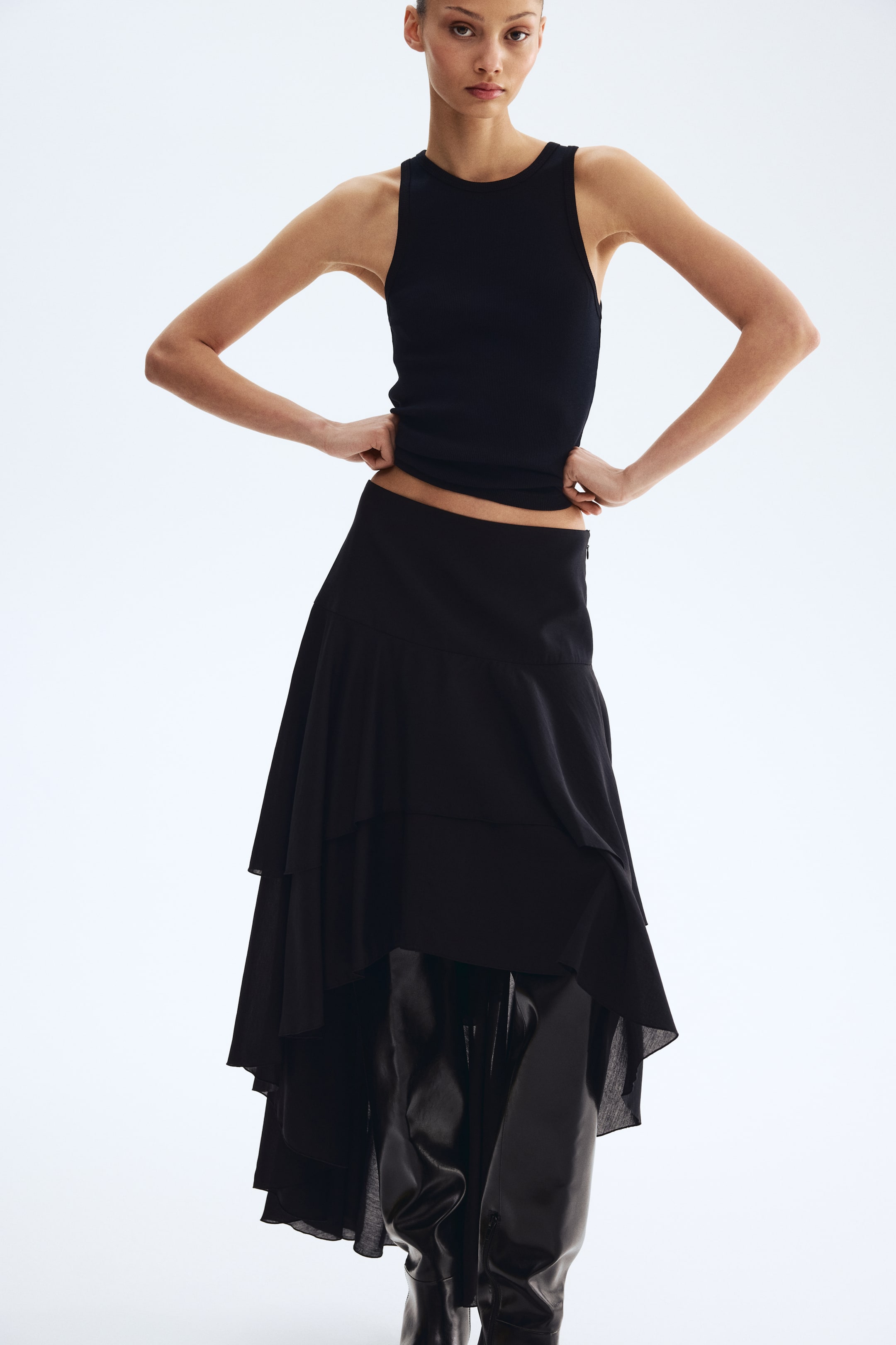 View larger image: The black ribbed tank top features a round neck and a fitted silhouette, with its hem slightly raised. The black skirt showcases an asymmetrical, tiered design with flowing fabric, partially revealing shiny black boots underneath.