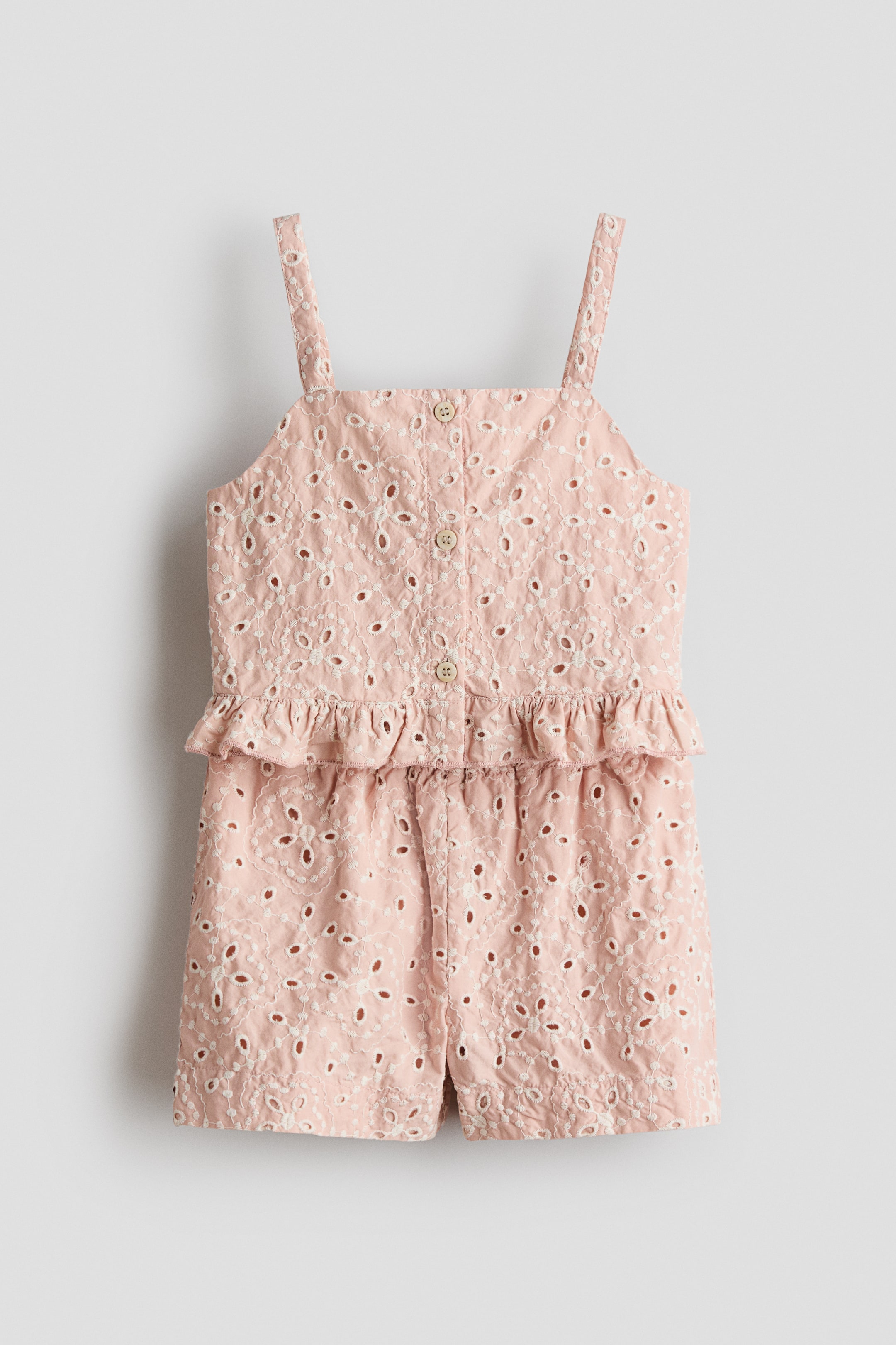 Ver imagen más grande: Un mono rosa presenta un patrón de encaje de ojales en todo su frente. Está diseñado con finos tirantes, una hilera de botones en la parte delantera con botones de color claro y un detalle de volante peplum en la cintura, que se extiende hacia los pantalones cortos.