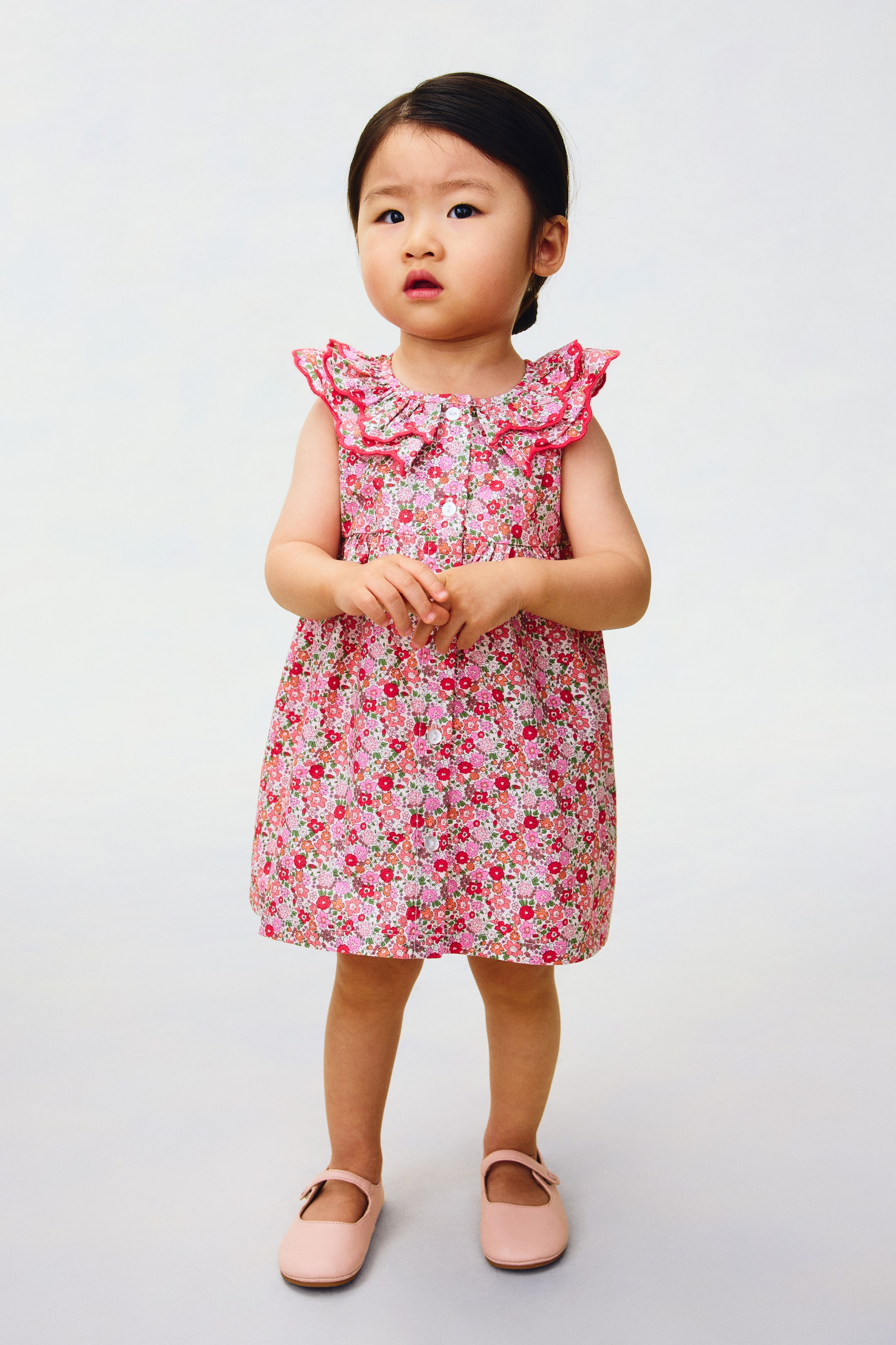 View larger image: A patterned dress showcases its front, featuring a floral print in shades of pink, red, green, and white. It has a round neckline, a double-layered scalloped collar with overlocked edges, and is sleeveless. Buttons run down the front, and a gathered seam above the waist shapes a flared skirt.