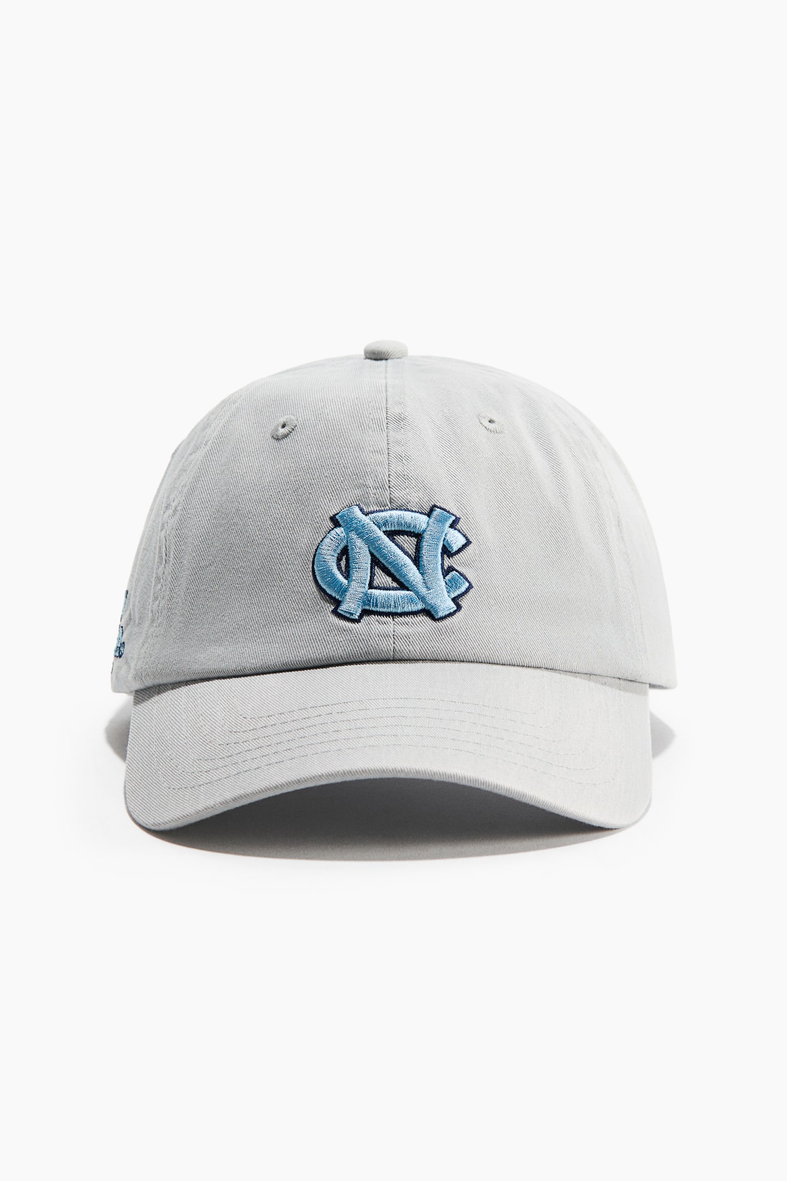 Un cappellino da baseball grigio chiaro Ã¨ rivolto frontalmente, con struttura a sei pannelli, visiera curva e bottone superiore. Sul davanti Ã¨ presente un logo "NC" intrecciato ricamato in azzurro chiaro con profilo blu scuro.