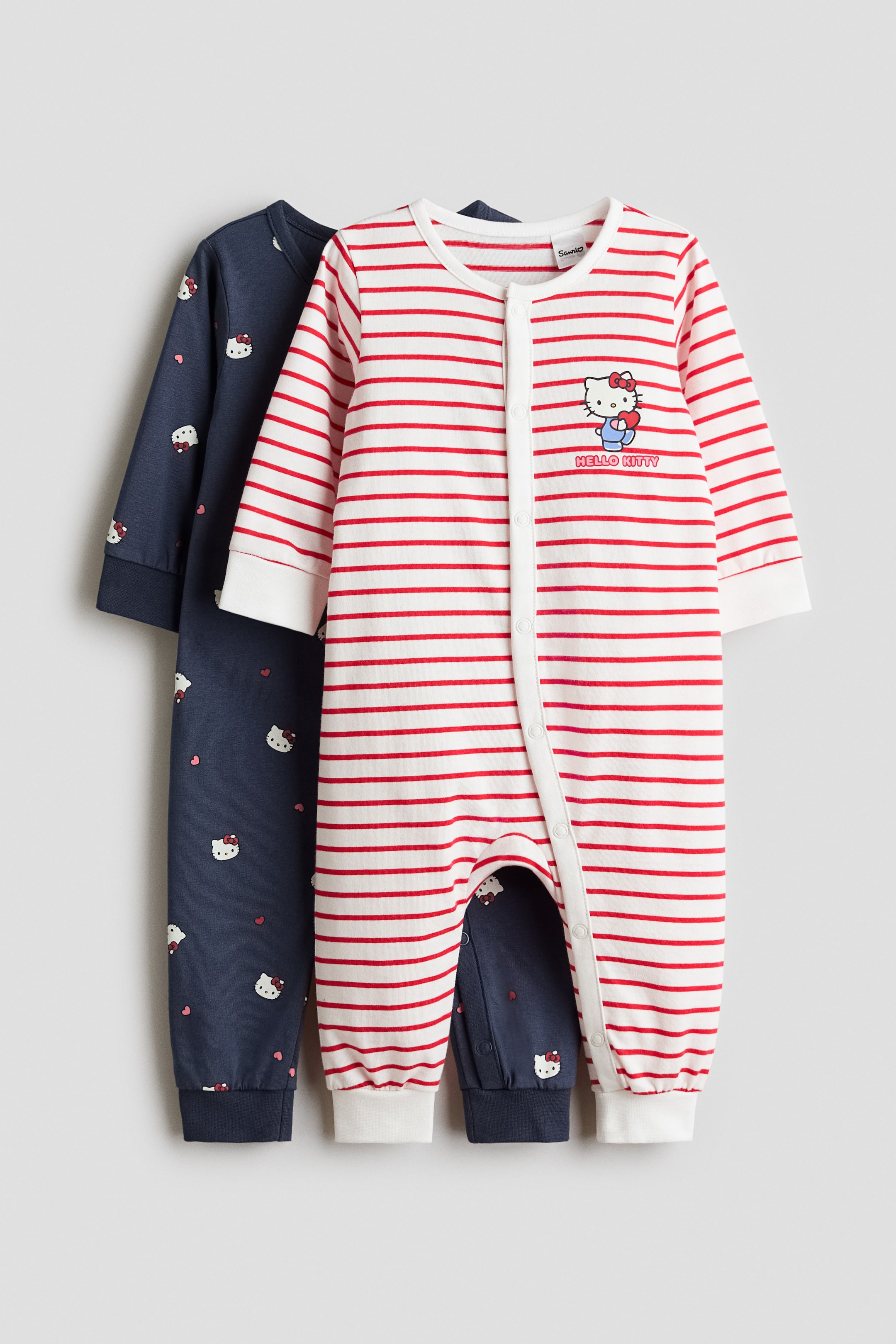 Visualizza immagine più grande: Due tutine per bebé sono presentate: una tutina bianca a righe orizzontali rosse, vista frontalmente, è sovrapposta a una tutina blu navy. La tutina a righe presenta una grafica di Hello Kitty sul petto e una chiusura con bottoni a pressione lungo tutta la lunghezza, mentre la tutina blu navy mostra una stampa all-over di volti di Hello Kitty e piccoli cuori.