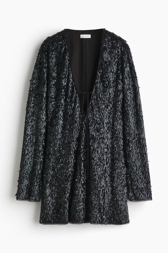 A black jacket is hanging, presenting its front, featuring an all-over texture of short, shimmering, fringe-like embellishments. It is designed with an open V-neckline, long sleeves, and thin ties are showing at the center front. - 1