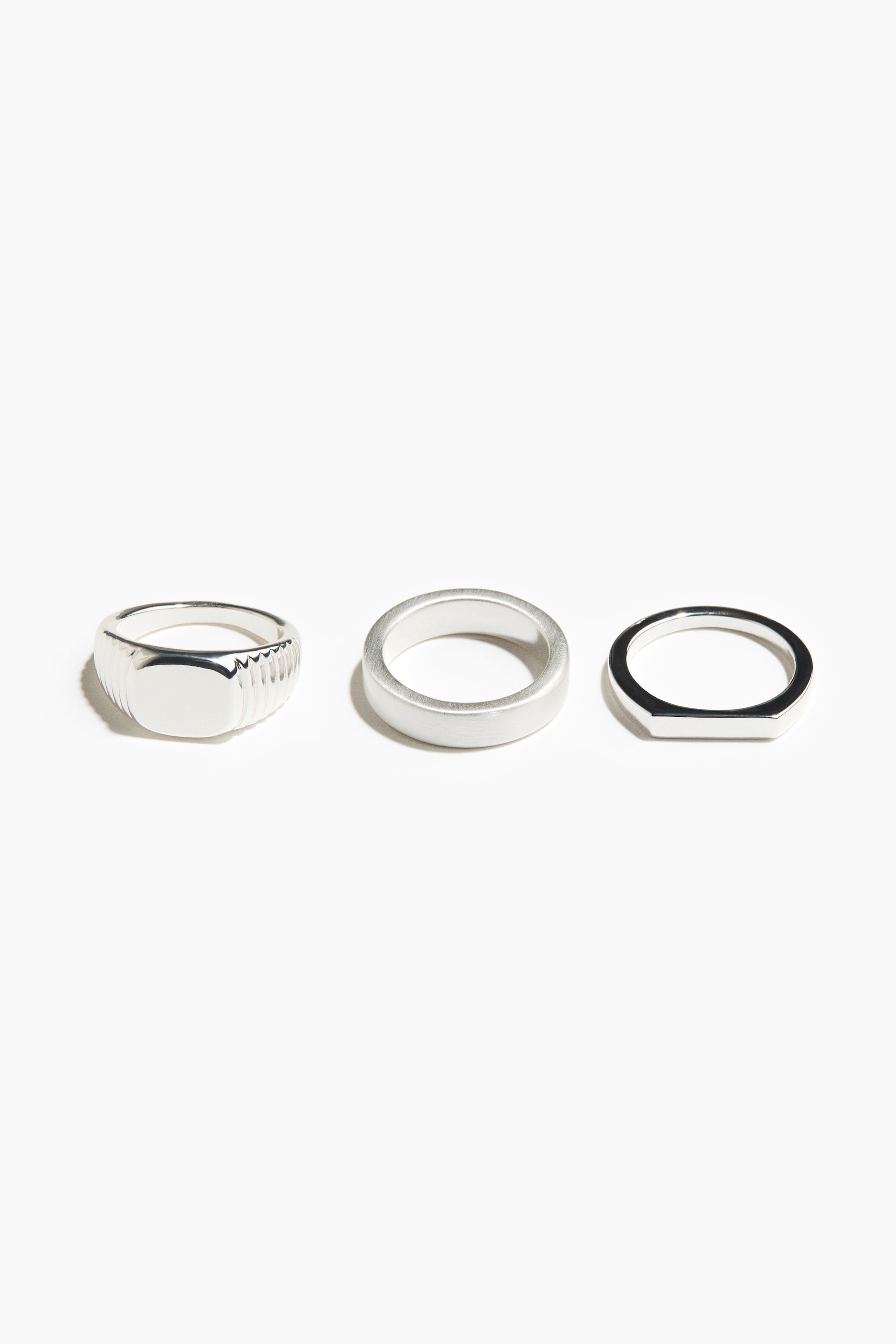 View larger image: Three silver-toned rings are showcased, each boasting a distinct design. The left ring features a polished signet style with a flat top and ribbed sides. The middle ring highlights a brushed, flat band, while the right ring presents a polished, thin band with an elongated flat top.