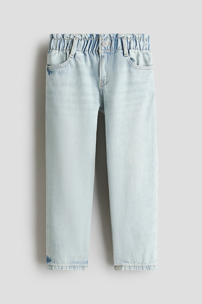 Un jean en denim de coton bleu clair est présenté de face, avec une taille en papier froissé élastiquée, une fausse braguette avec un bouton-pression et des poches latérales en diagonale. Ils sont conçus avec une coupe ample au niveau des hanches et des cuisses, s'affinant doucement le long des jambes. - 1