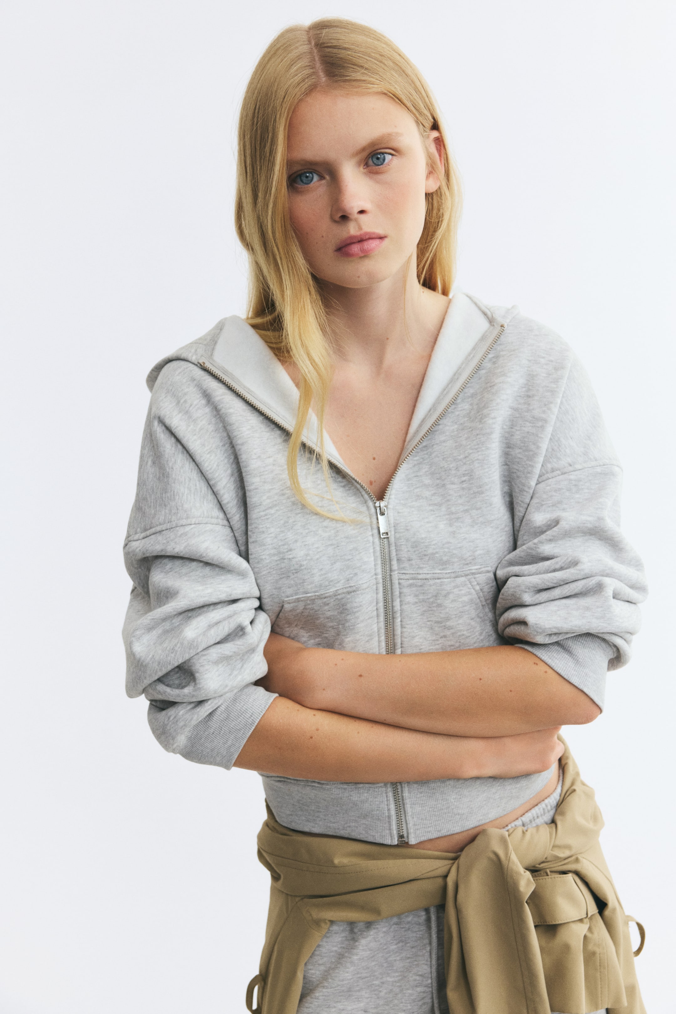View larger image: A light grey zip-up hoodie in sweat fabric is worn by the model, who is posing with crossed arms. The hoodie features a partially unzipped front revealing a light-coloured lining, dropped shoulders, and ribbed cuffs, and is styled with matching grey sweatpants and a beige garment tied around the waist.