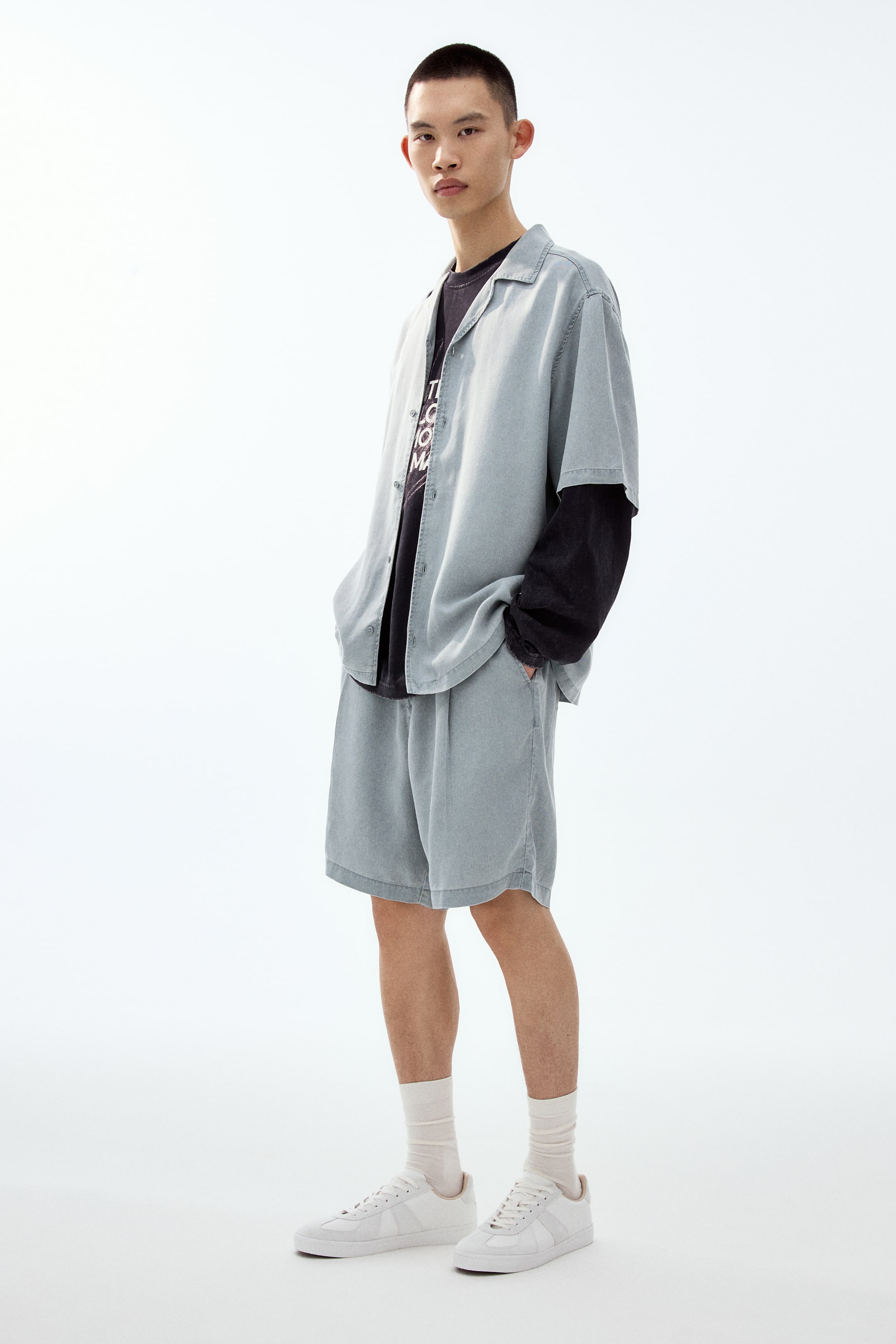 Xem ảnh lớn hơn: Bộ áo tay ngắn màu xanh nhạt và quần short đồng bộ đang được trưng bày, hướng về phía trước. Áo có cổ camp, khoác hờ ngoài một áo thun tay dài tối màu, còn quần short có phom suông thoải mái và túi hai bên nhìn thấy rõ.