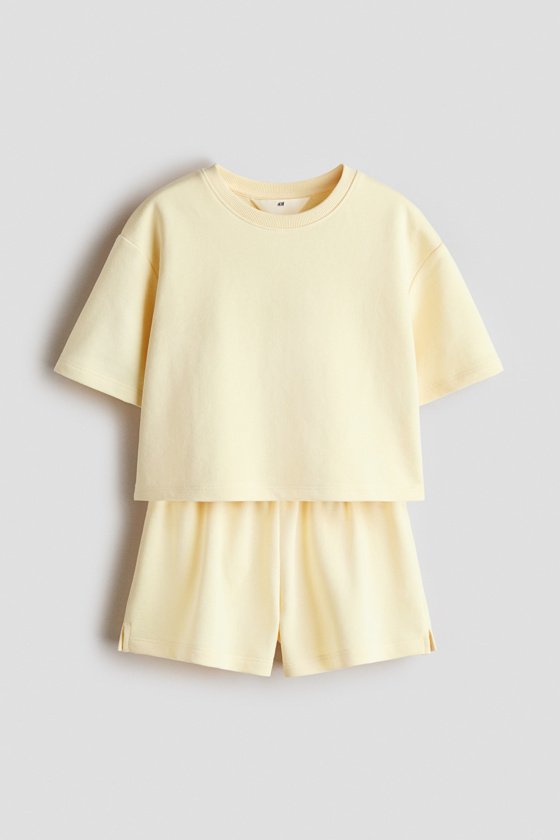 View larger image: A light yellow two-piece set, crafted from lightweight sweatshirt fabric, is displayed from the front, showcasing a T-shirt with a round, rib-trimmed neckline, dropped shoulders, and a straight hem with short side slits. The matching shorts are designed with covered elastication at the waist and short slits at the hem.