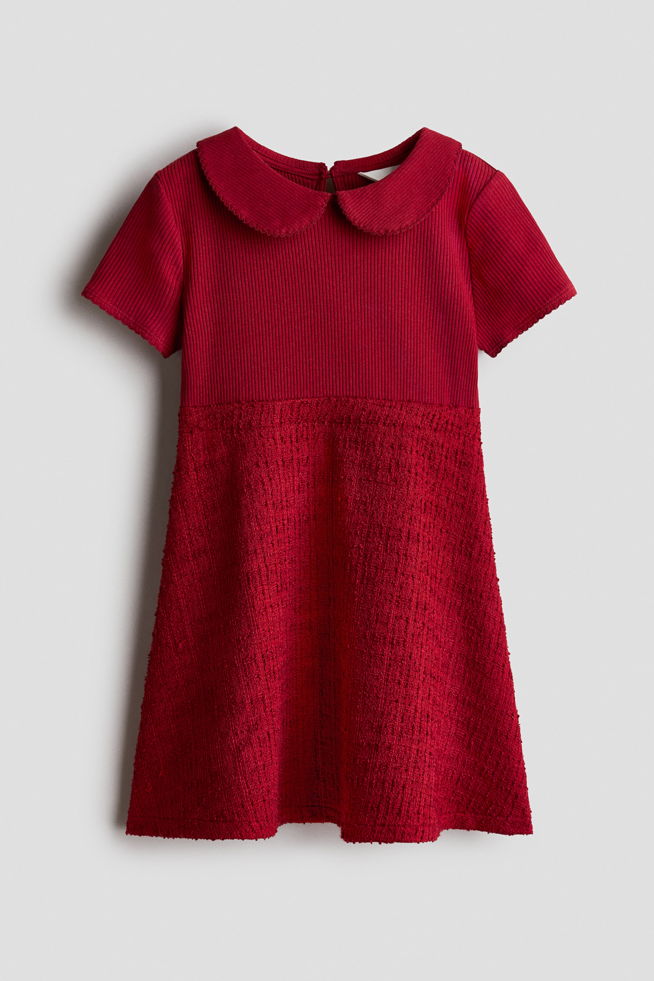 View larger image: A red flared skirt collared dress is shown from the front, featuring a ribbed-knit bodice with a rounded Peter Pan collar and short sleeves. This bodice joins a textured, woven flared skirt that drapes downward.