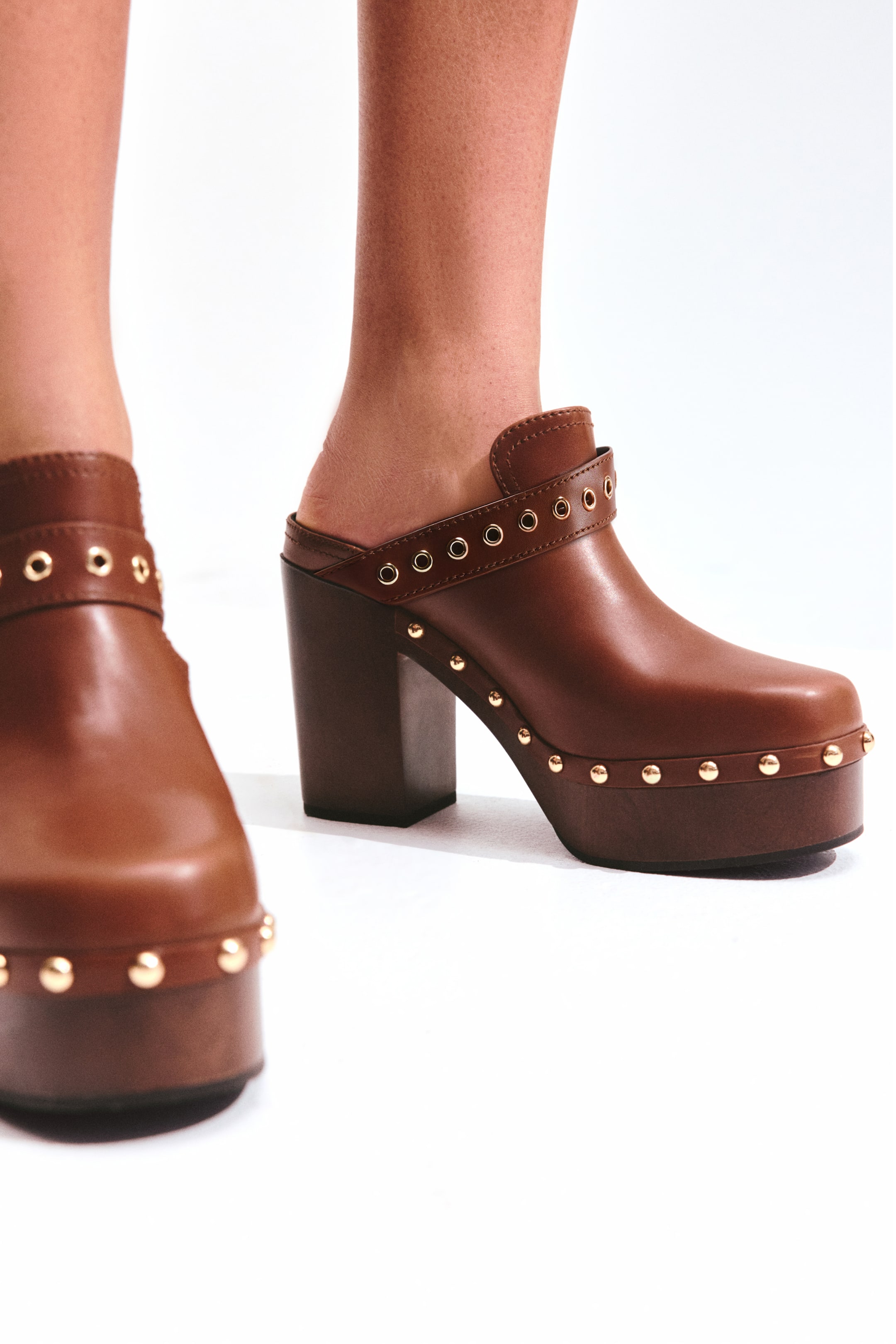 View larger image: Brown leather platform clogs feature a rounded toe, a chunky block heel, and a thick platform sole embellished with gold-toned studs. Gold-toned eyelets detail a strap across the instep, as a foot models the open-back clog from a three-quarter side view.
