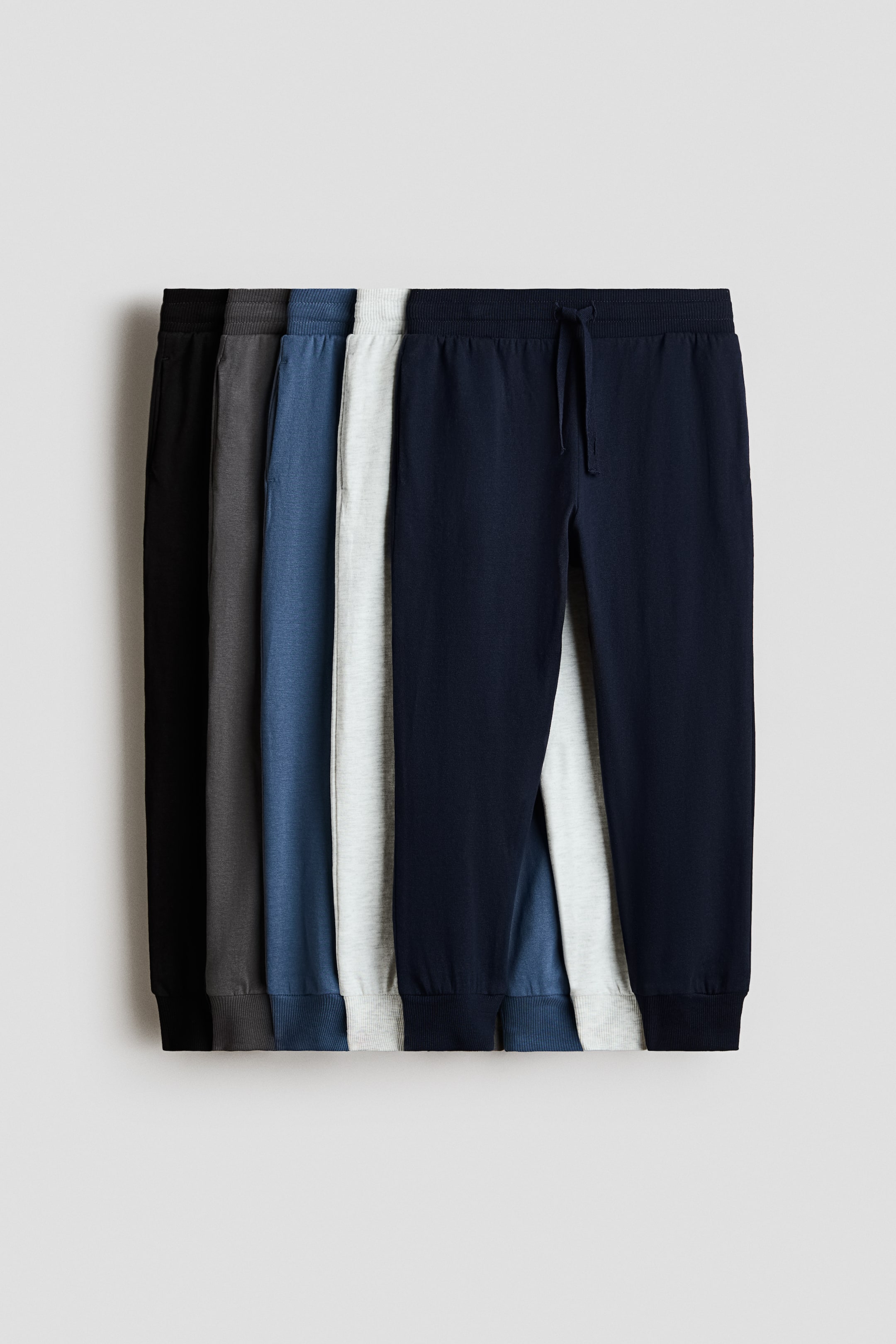 Größeres Bild ansehen: Fünf Paar Jogginghosen sind übereinandergelegt und leicht versetzt, um ihre Vorderansicht zu präsentieren. Jedes Paar ist mit einem elastischen Bund mit Kordelzug und gerippten Bündchen ausgestattet und erscheint in den Farben Schwarz, Dunkelgrau, Blau, Hellgrau und Marine.