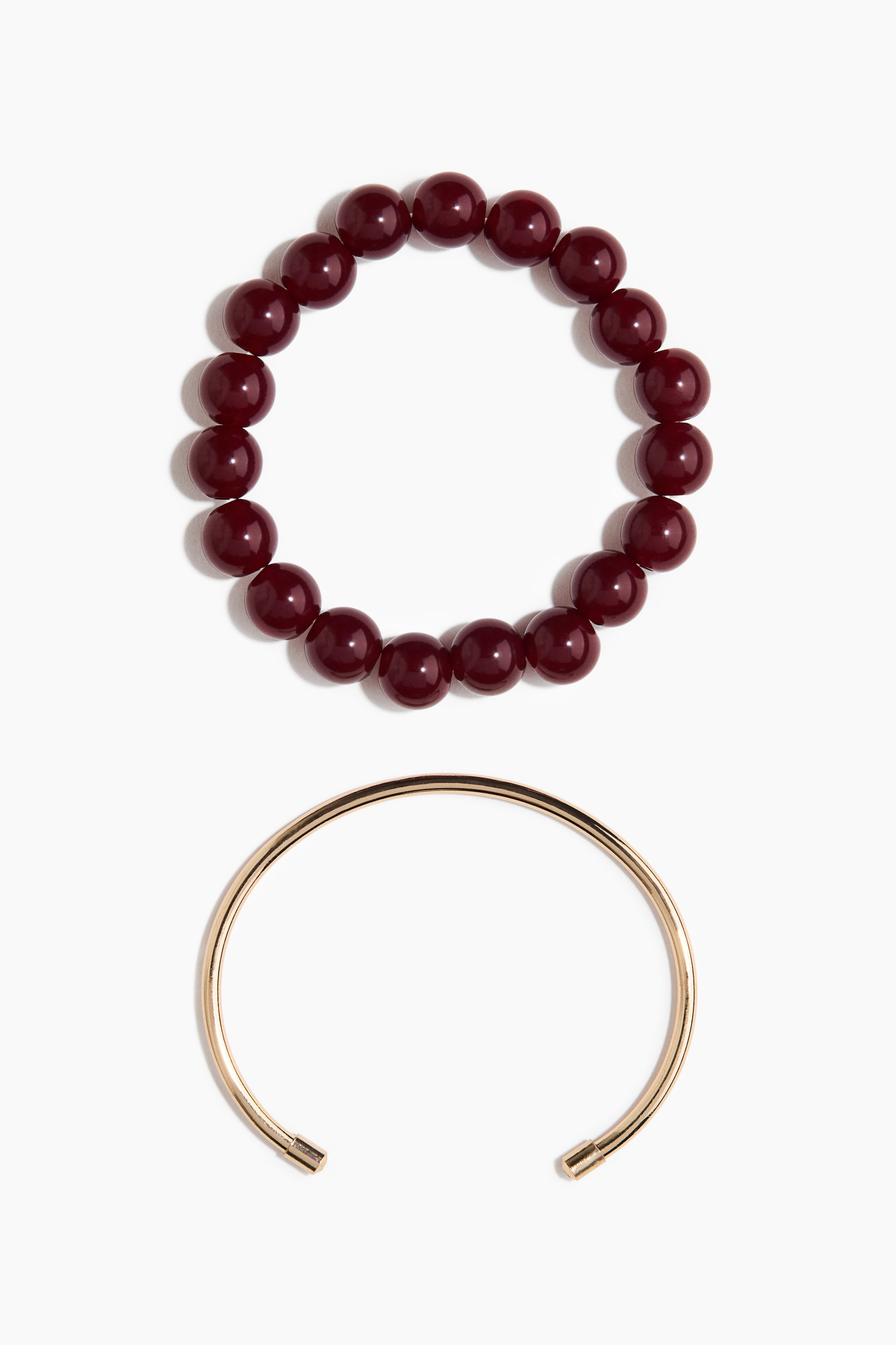 Grotere afbeelding bekijken: Een set van twee armbanden ligt plat, met een bovenarmband met glanzende, donkerrode ronde kralen en een onderarmband die een gladde, goudkleurige open manchet is met kleine cilindrische dopjes aan elk uiteinde.