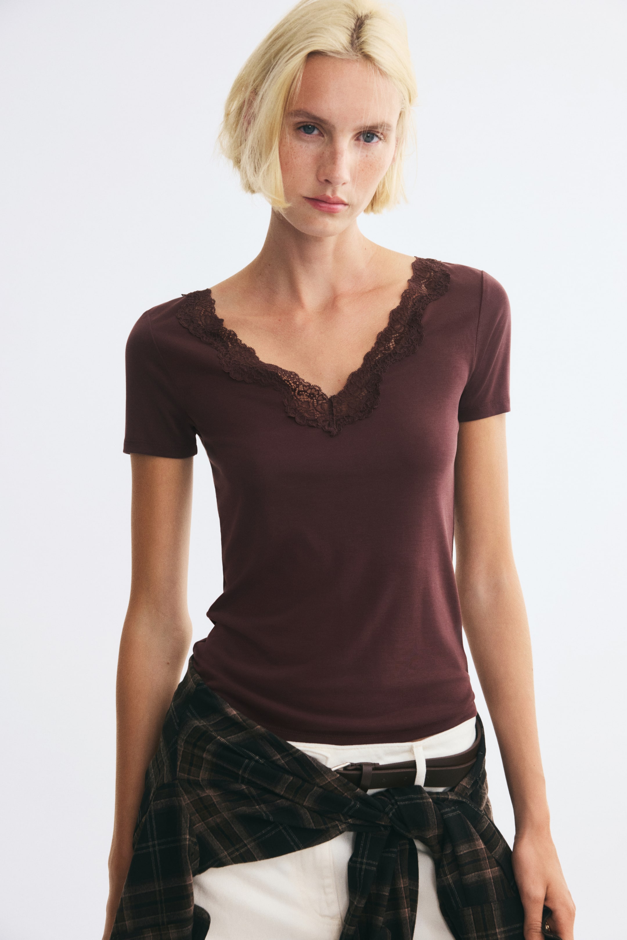 View larger image: A fitted dark brown modal-blend jersey top with a deep V-neck trimmed with lace and short sleeves is worn by the model. The top is styled with white trousers and a plaid shirt tied around the waist.