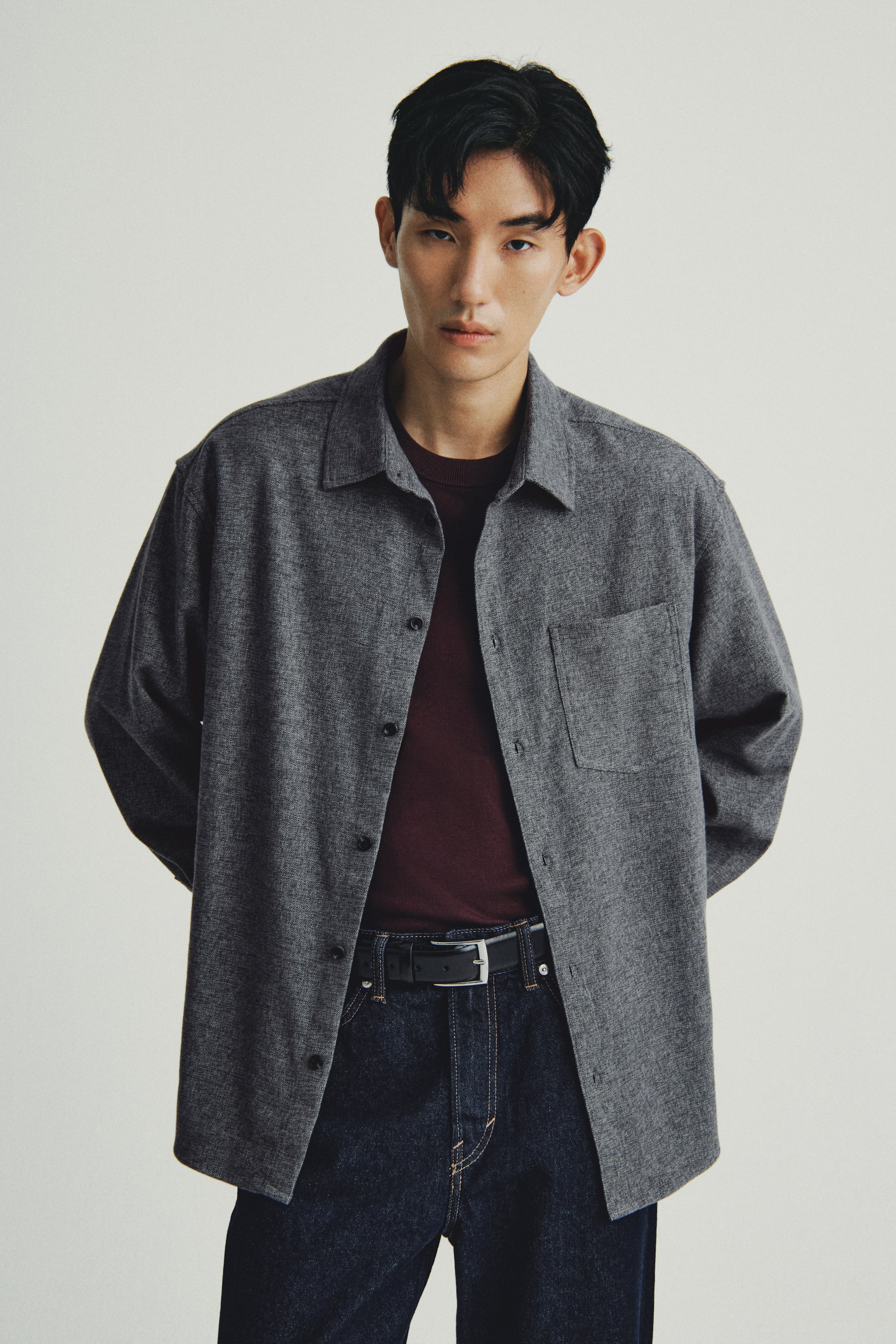 이미지 확대 보기: 회색 혼방 셔츠가 앞을 향하고 있으며, 뾰족한 칼라와 앞쪽 플라켓을 따라 달린 어두운 버튼, 왼쪽 가슴 위의 단일 패치 포켓이 특징입니다. 셔츠는 어두운 빨간색 긴팔 티셔츠 위에 열려 있으며, 여유로운 핏을 자랑하고 있습니다.