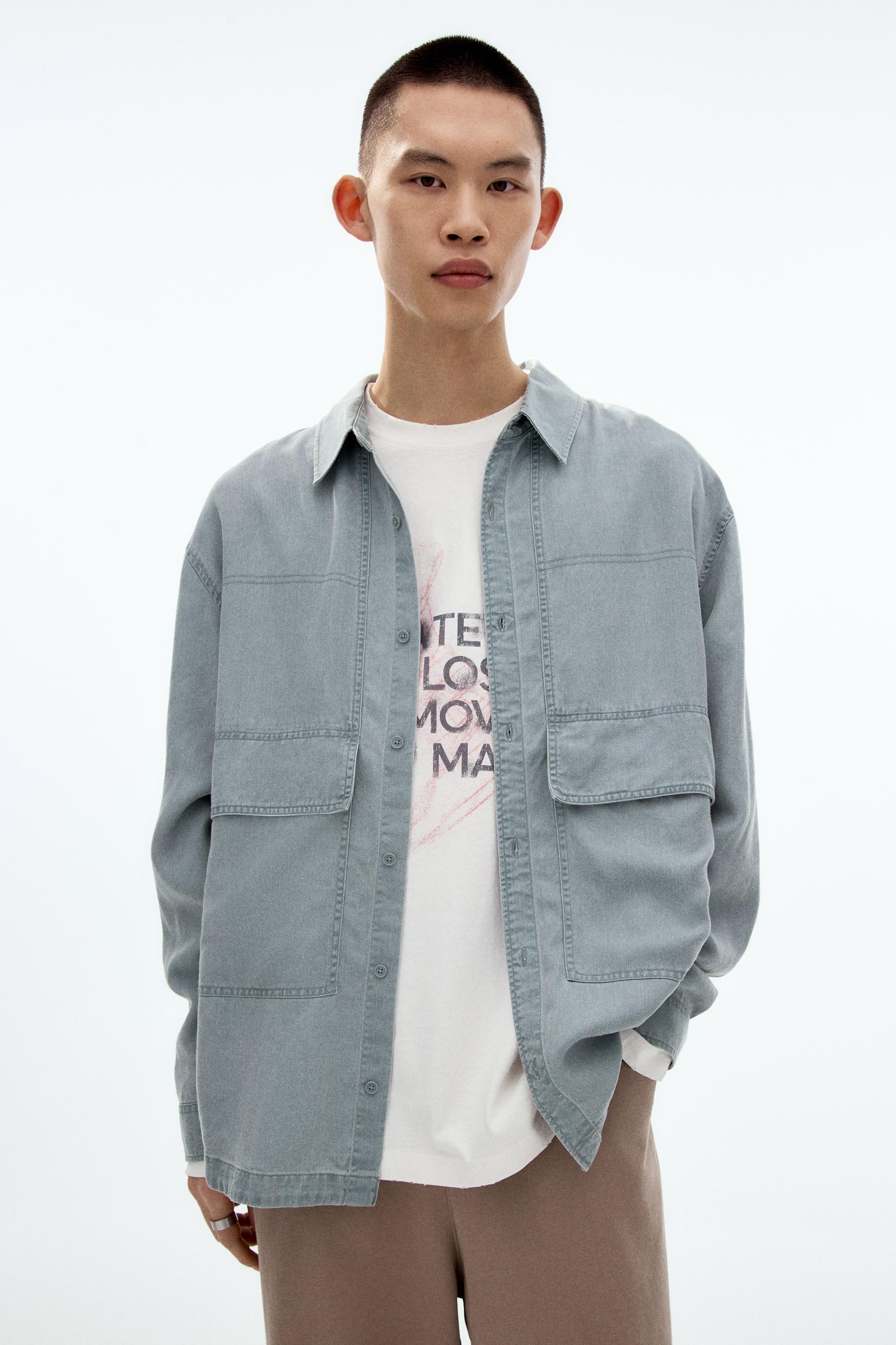 View larger image: A light blue-grey overshirt features a pointed collar, a button-front fastening, two chest patch pockets with flaps and two lower patch pockets. It is worn open over a white T-shirt, showcasing visible stitching details and a relaxed fit.