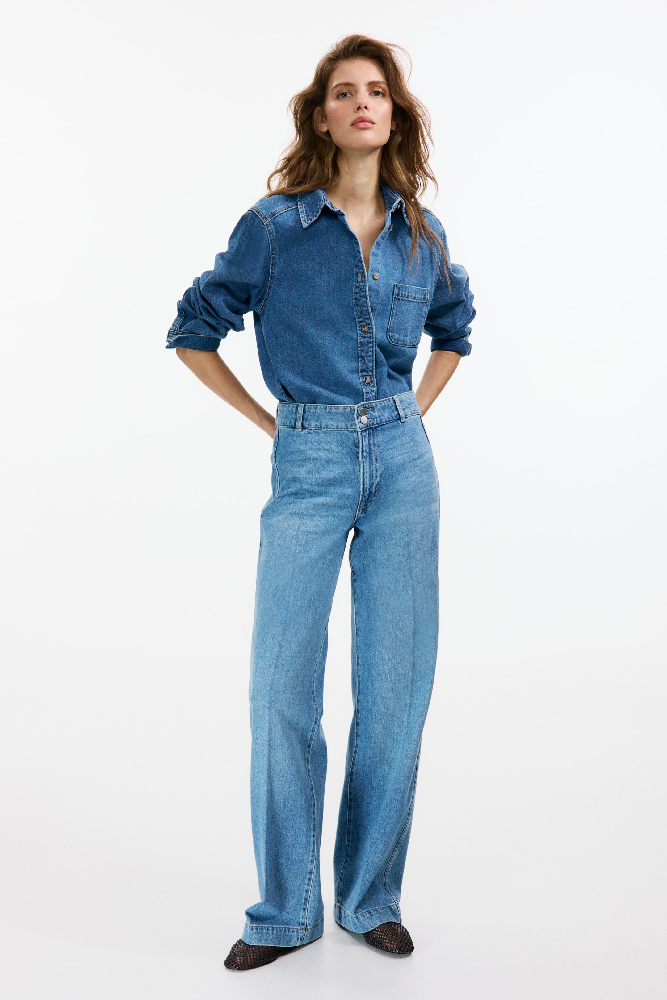 View larger image: The model is wearing light blue wide-leg jeans with a high waist, featuring a zip fly and two buttons. These jeans showcase creases along the front and back, extending down to the top of the foot. They are styled with a darker denim shirt tucked in and paired with mesh shoes.