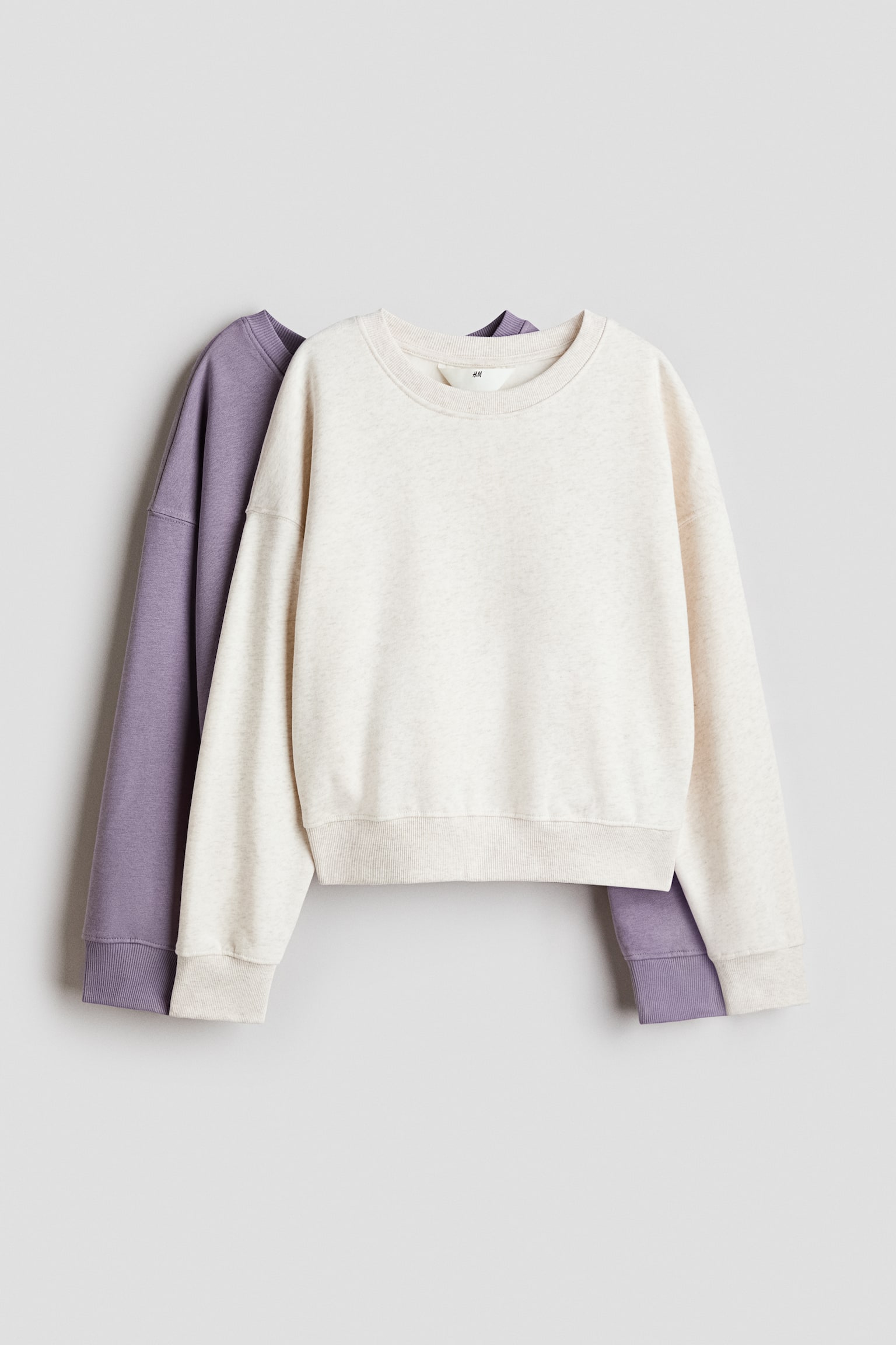 Two loose-fit jumpers in a brushed fleece fabric are hanging facing forwards, with a light-beige jumper layered directly over a purple one. Both garments feature a round, rib-trimmed neckline, dropped shoulders, and ribbing at the cuffs and hem.