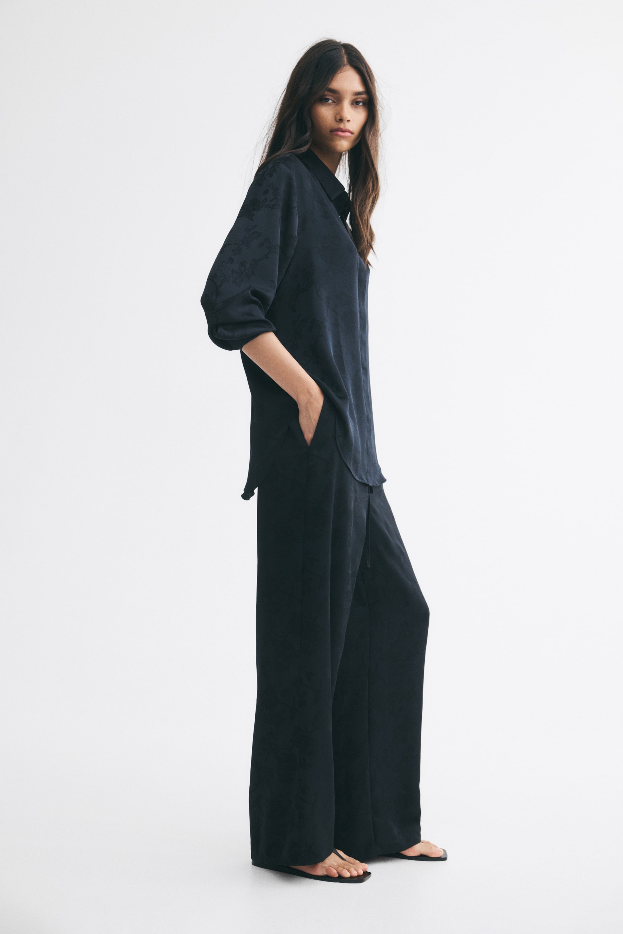 View larger image: The model is wearing a dark navy jacquard shirt with a subtle floral pattern, paired with matching wide-leg trousers. The shirt features a collar, long sleeves, and a curved hem, with the model standing in profile and resting one hand in the trouser pocket.
