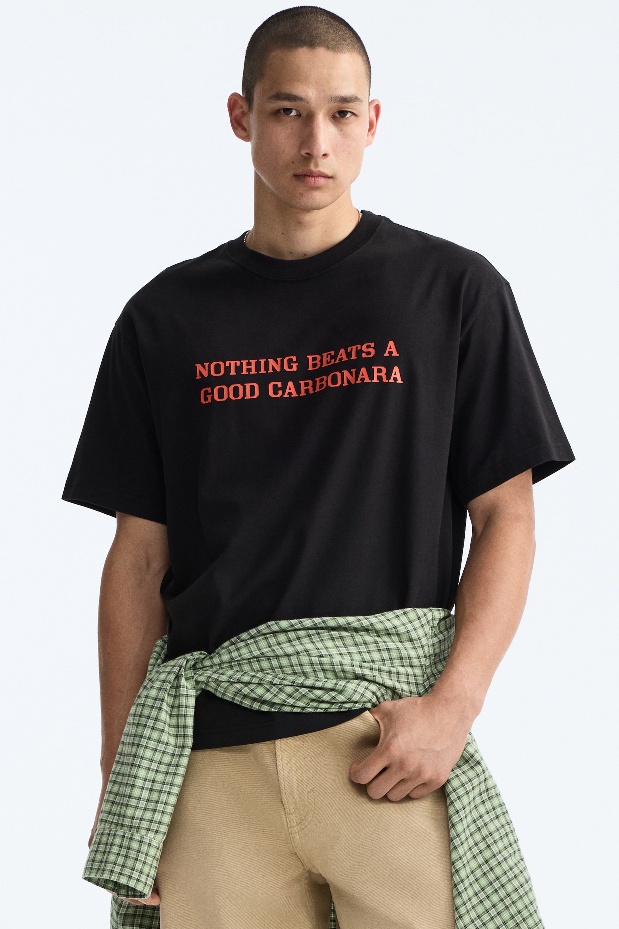 View larger image: The model is wearing a black T-shirt adorned with a red print motif stating "NOTHING BEATS A GOOD CARBONARA." This T-shirt boasts a round, ribbed neckline, dropped shoulders, and a straight-cut hem, offering a relaxed fit. Additionally, a green plaid shirt is stylishly tied around the model's waist, complementing the T-shirt.