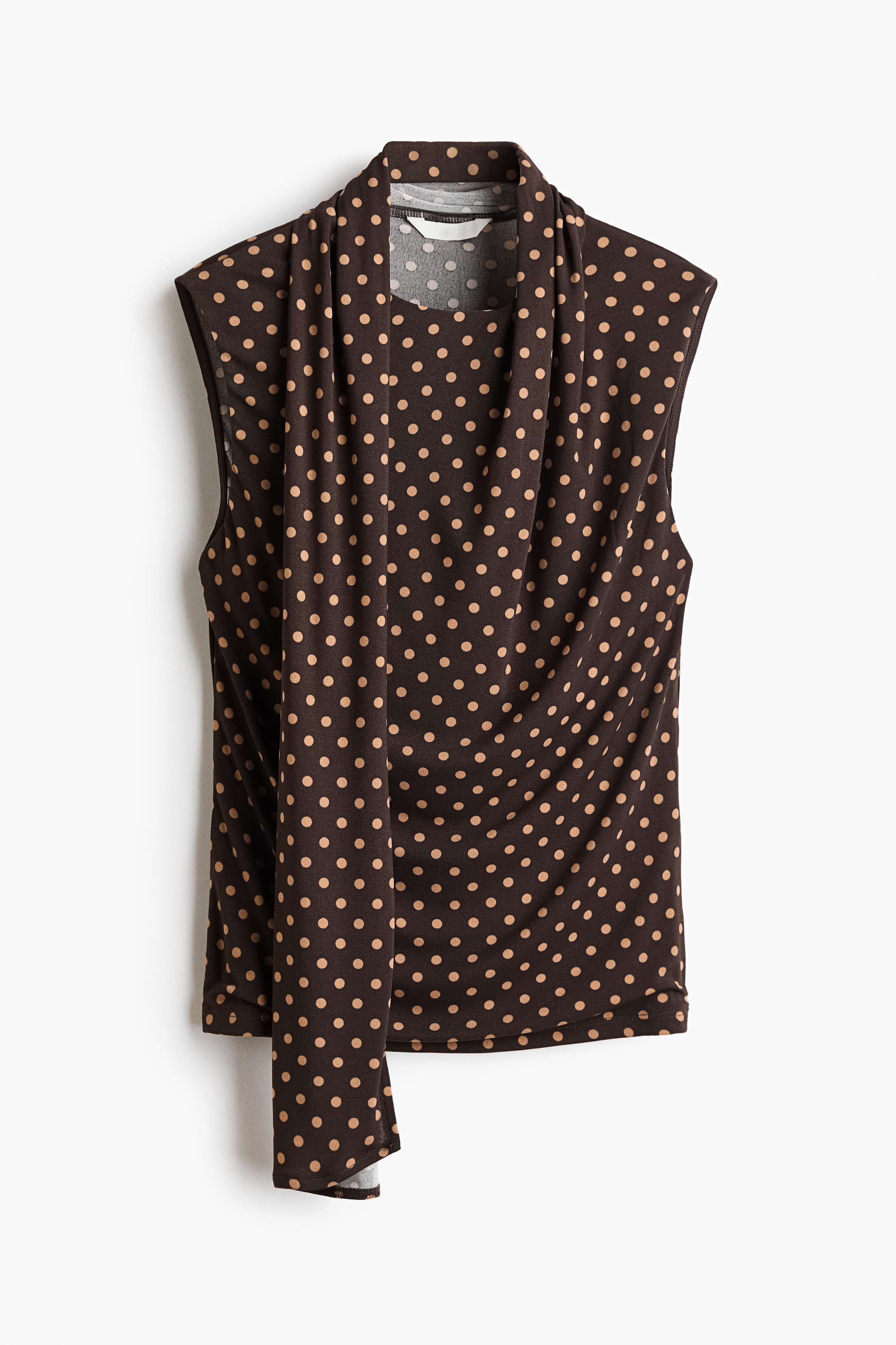 The model is wearing a sleeveless brown and beige polka-dot top, featuring a high collar with a matching scarf draping down the centre front. The top is styled with wide-leg black jeans.