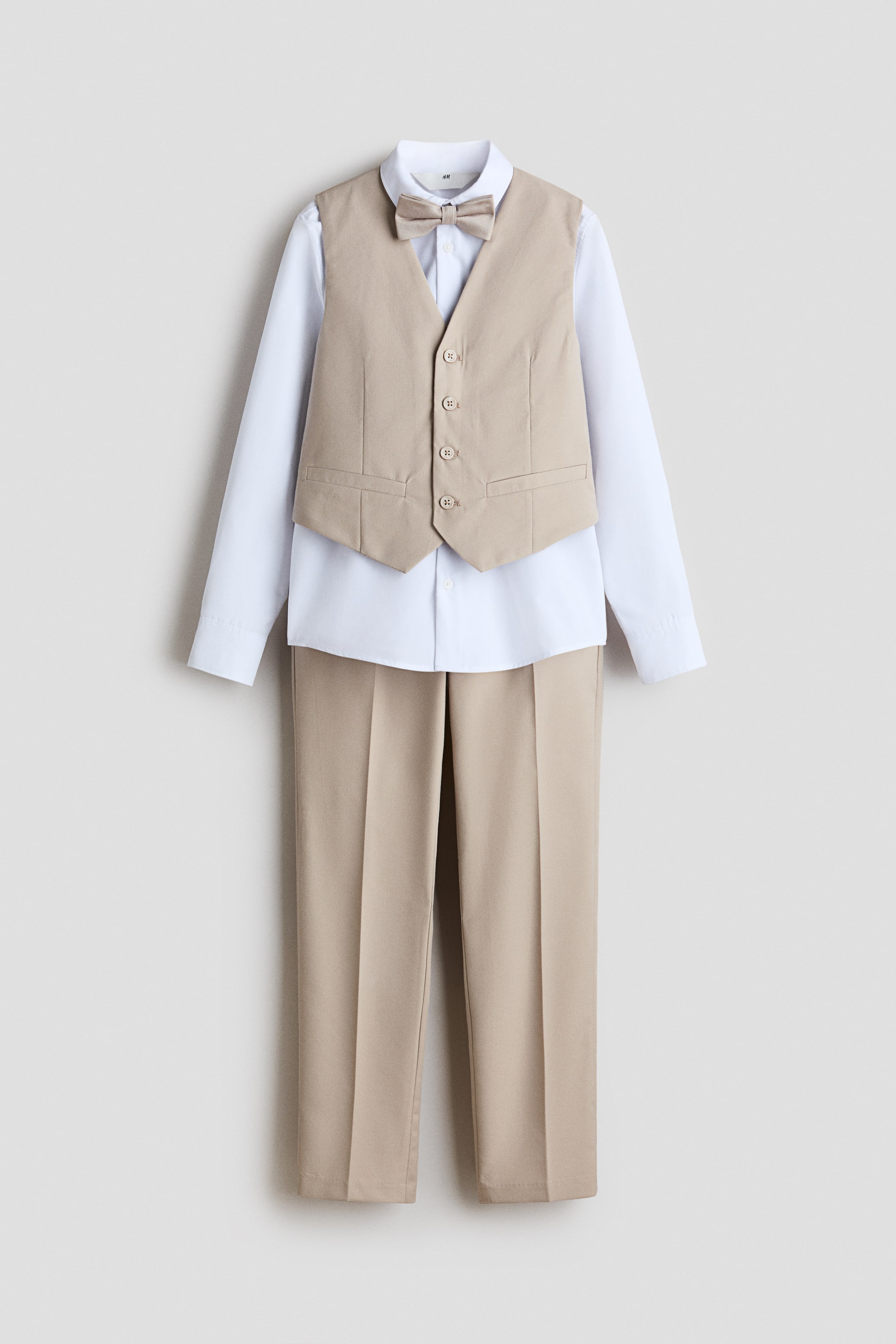 Visualizza immagine più grande: Un completo per bambini presenta una camicia bianca a maniche lunghe, stratificata sotto un gilet beige, pantaloni beige coordinati e un papillon beige. Il gilet, presentato FRONTALE, è caratterizzato da quattro bottoni e due tasche finte.
